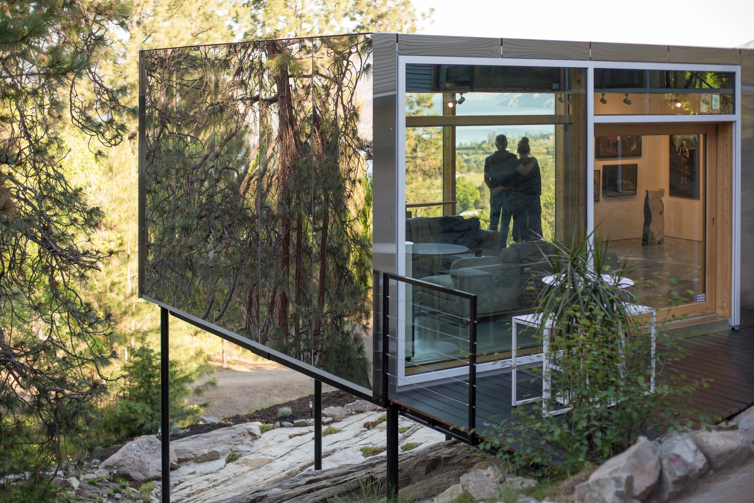 A modern house with a glass wall overlooking a forested landscape. Inside, two people stand near the window, looking out at the view.