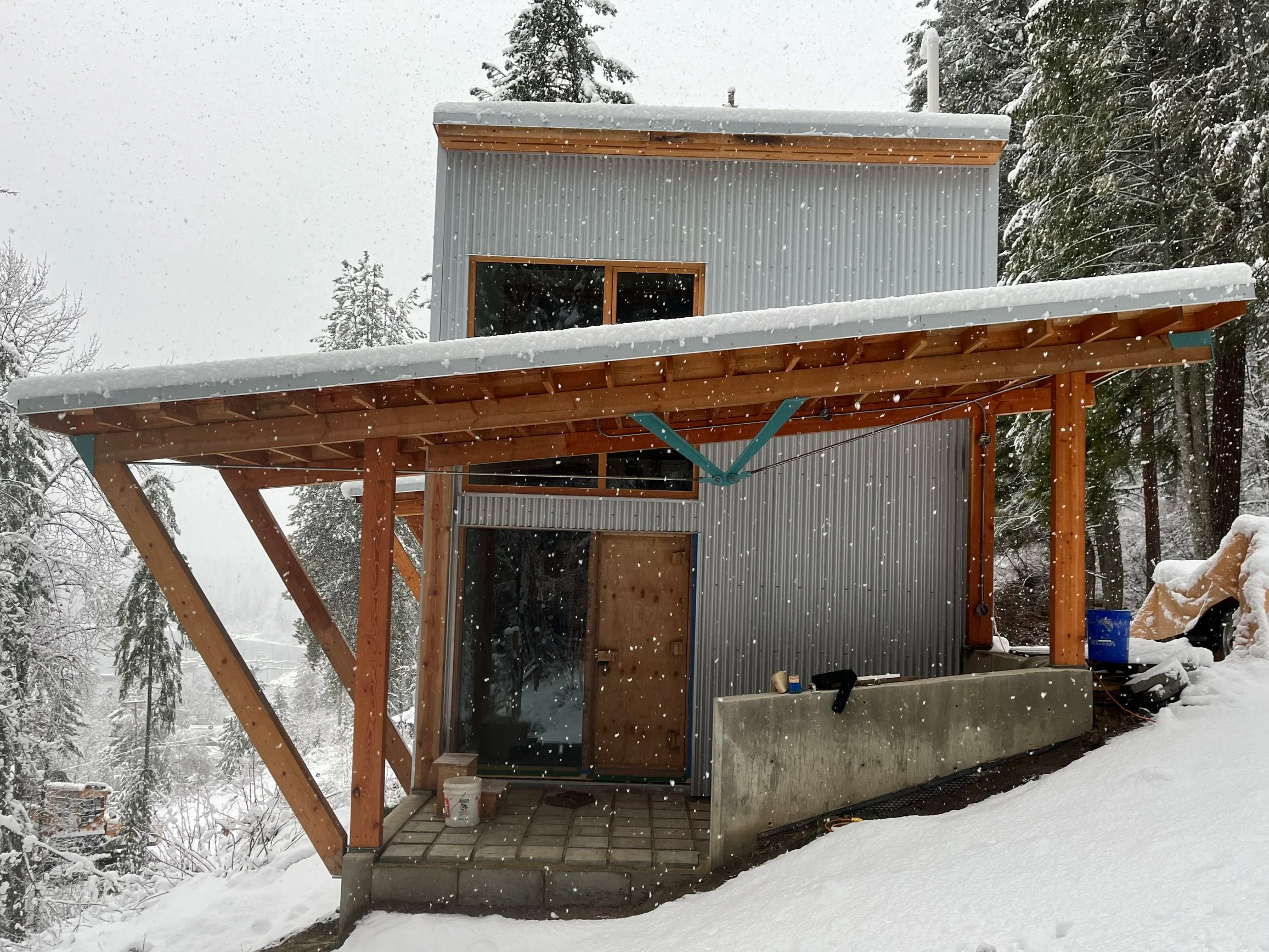 A small modern house with a metal exterior, wooden accents, and a snow-covered roof, located in a snowy, forested area.