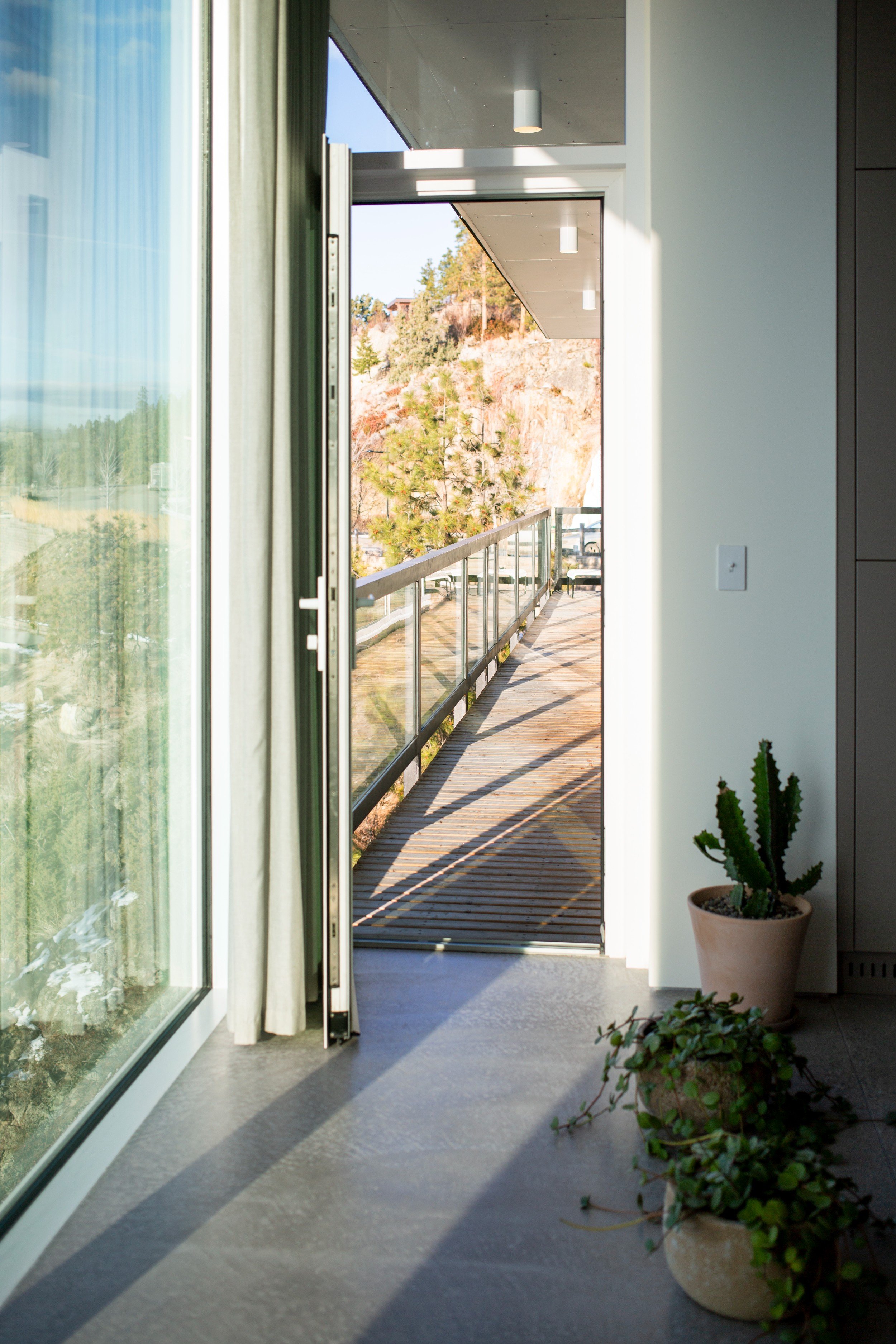 View of a modern balcony with a glass railing, wooden floor, and outdoor scenery of trees and rocky hillside, seen through an open door from the indoor area with potted plants.