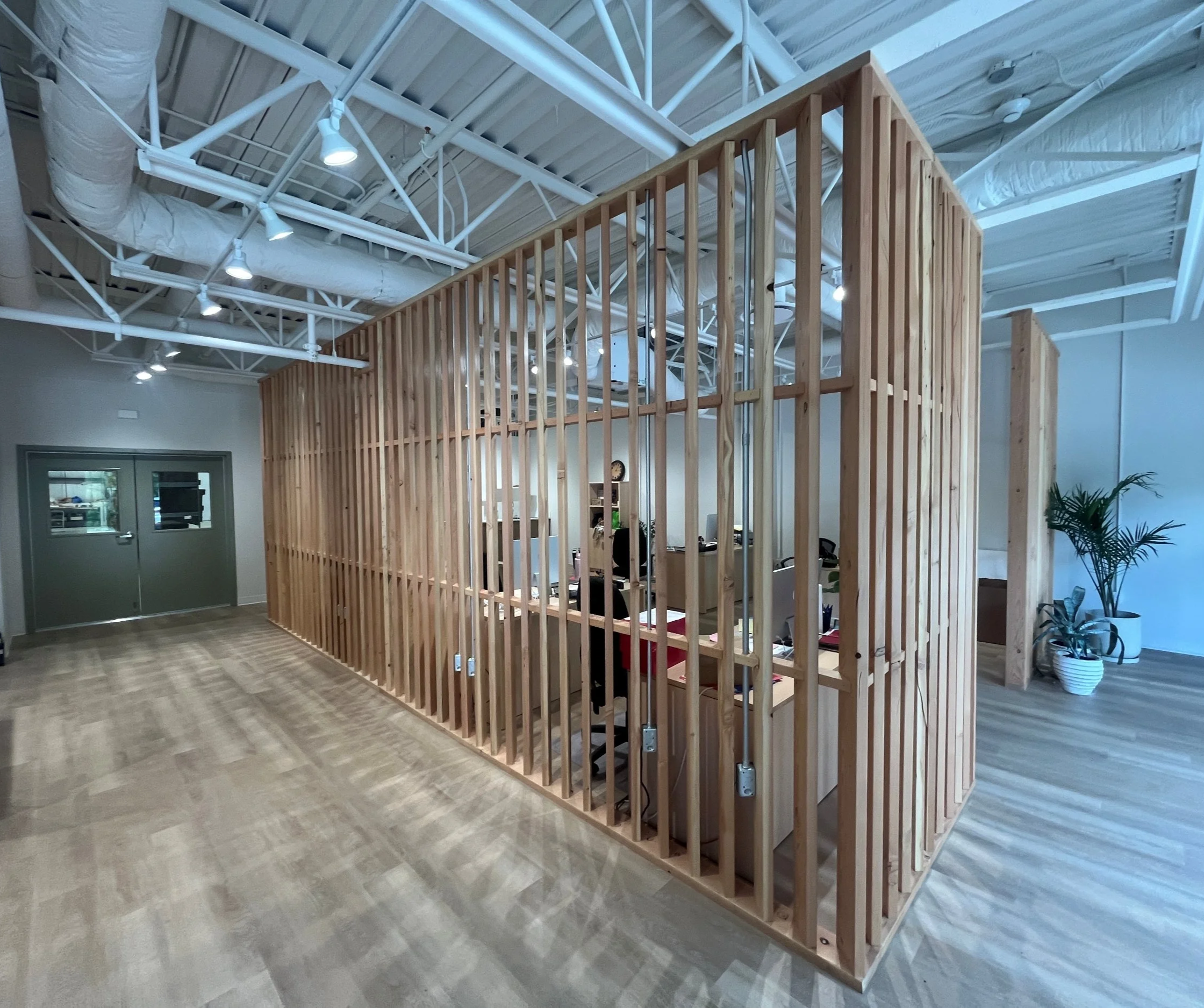 Interior view of an office or workspace with a wooden partition wall and visible ceiling ductwork, pipes, and lighting fixtures.