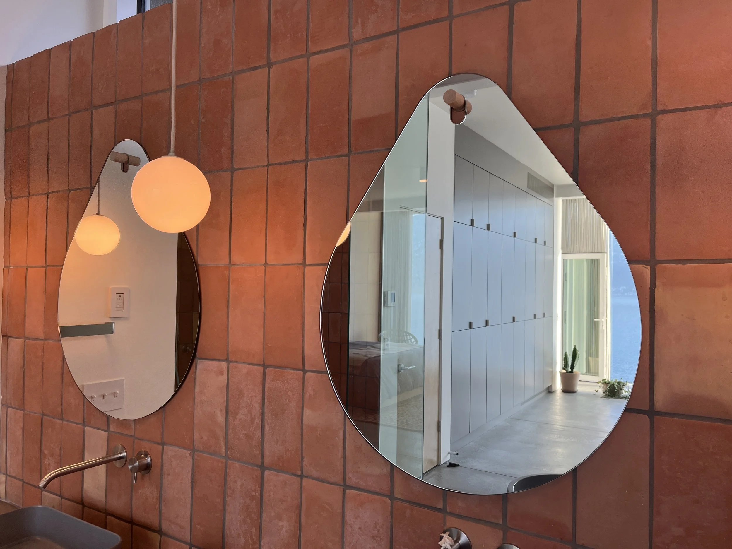Two irregularly shaped mirrors mounted on a terracotta tiled wall in a modern interior space, with a view of a bright room with white cabinets, a potted cactus plant, and a glass door leading to the outdoors.