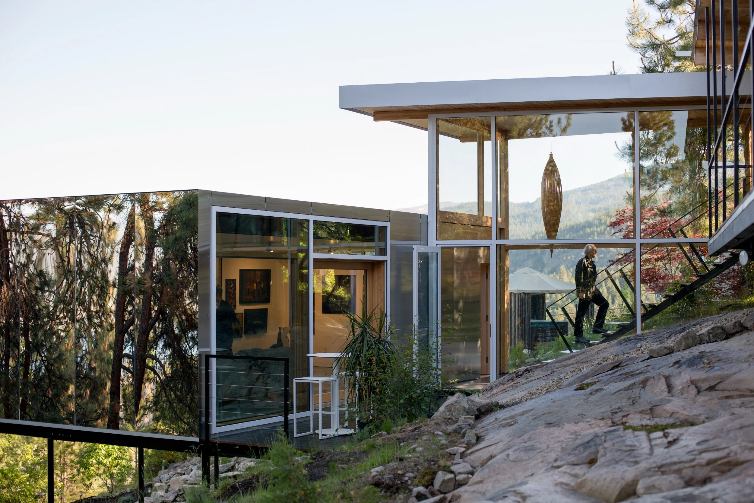 Modern glass house built on a rocky hillside overlooking trees and mountains, with a person walking up stairs outside.