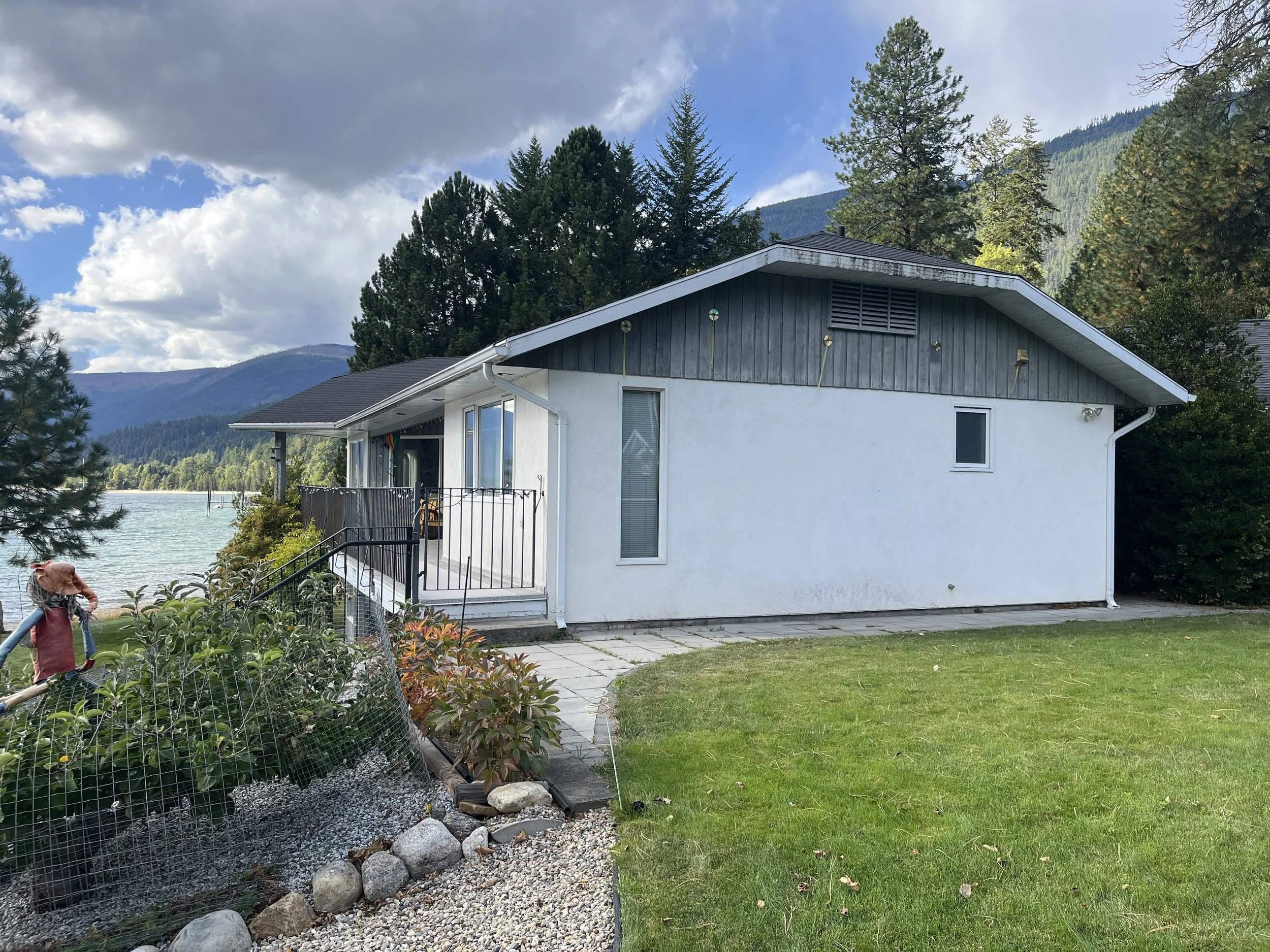 A house with a white exterior and dark wooden accents, situated near a lake, with surrounding trees and mountains in the background. There is a small porch with railing, paved pathway, and a grassy lawn.