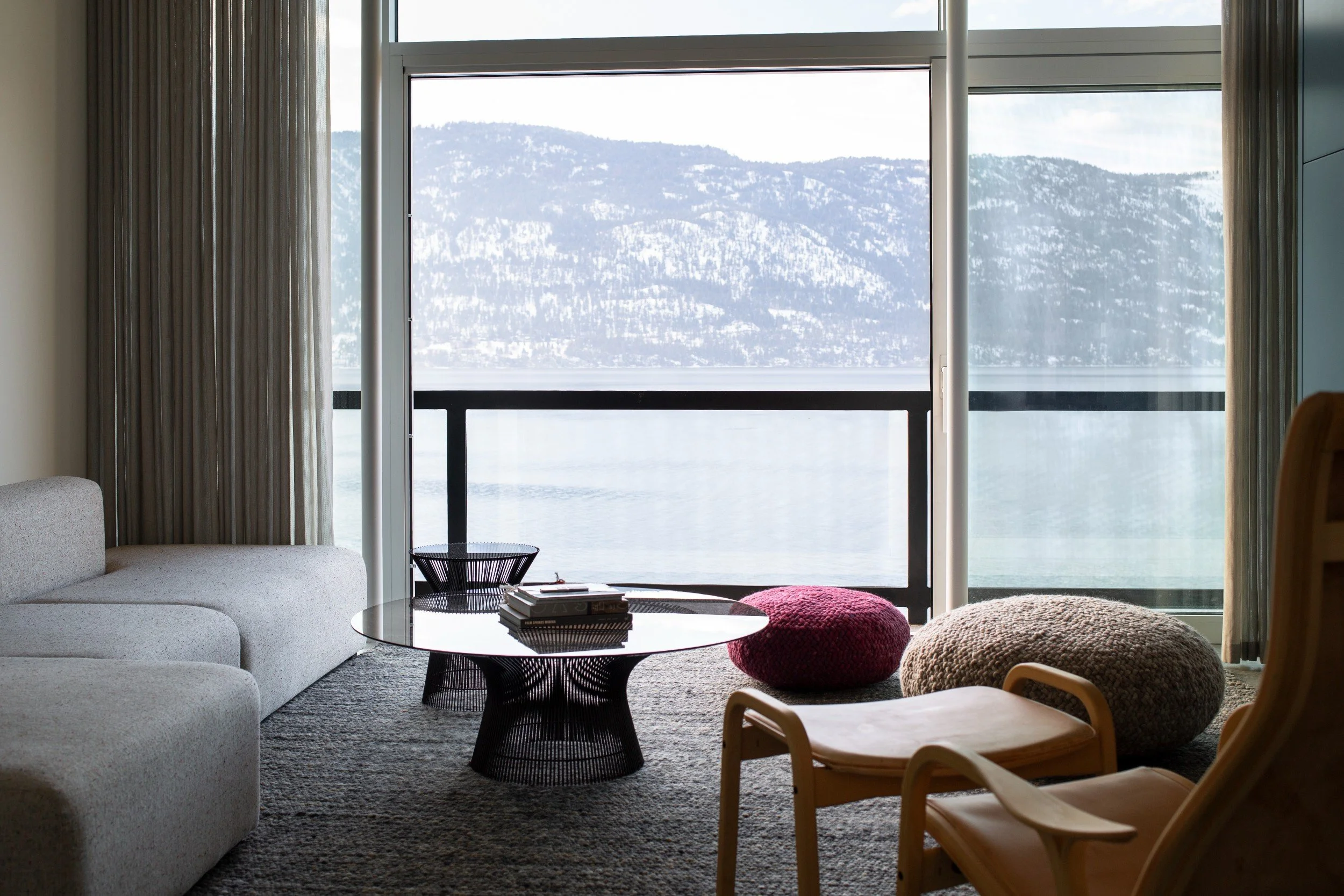 Living room with large glass window overlooking snowy mountains and lake, with sofa, coffee table, and colorful floor cushions.