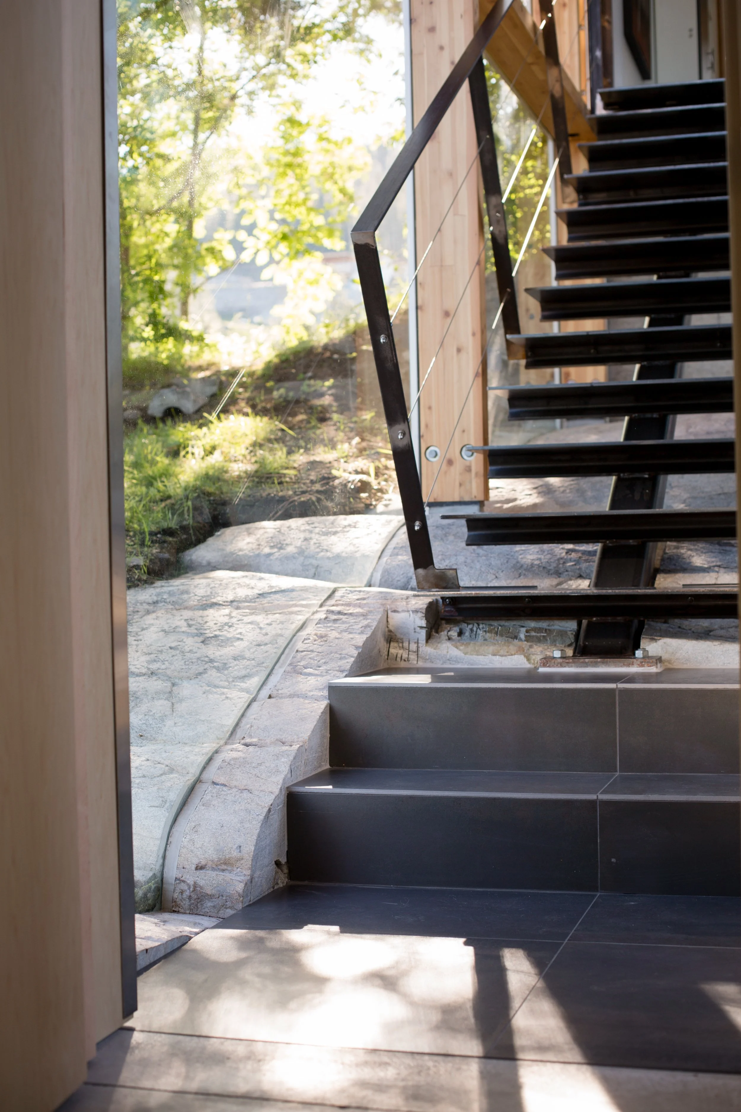 Interior view of a staircase with black steps, metal railing, and large window showing green trees outside, with sunlight streaming in.