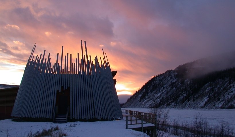 A round wooden building with vertical logs and an open doorway, set against a snowy landscape with mountains and a colorful sunset sky.