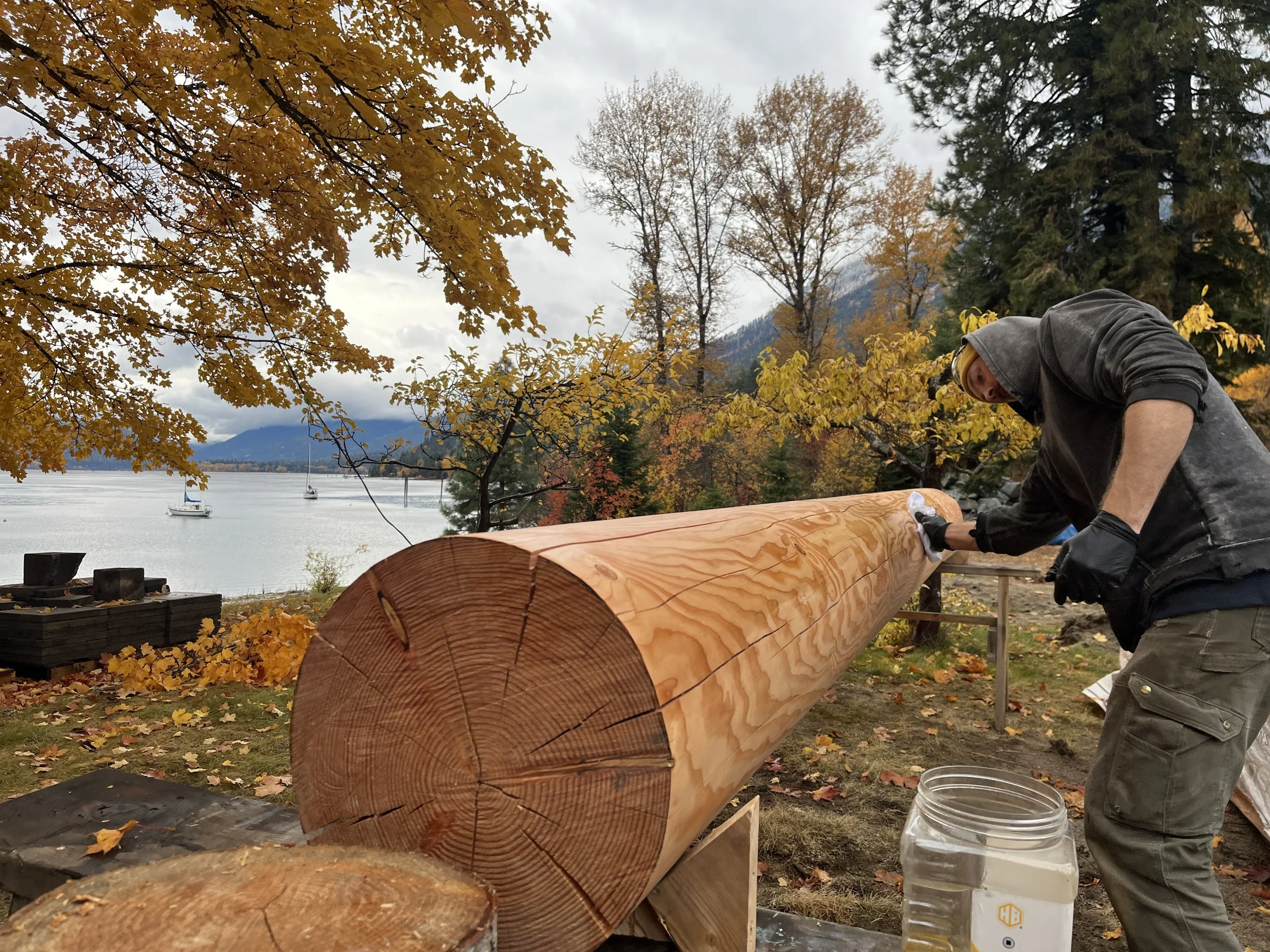 A person in a hoodie and gloves is carving or sanding a large log outdoors near a lake with sailboats and mountains in the background, surrounded by colorful autumn trees.