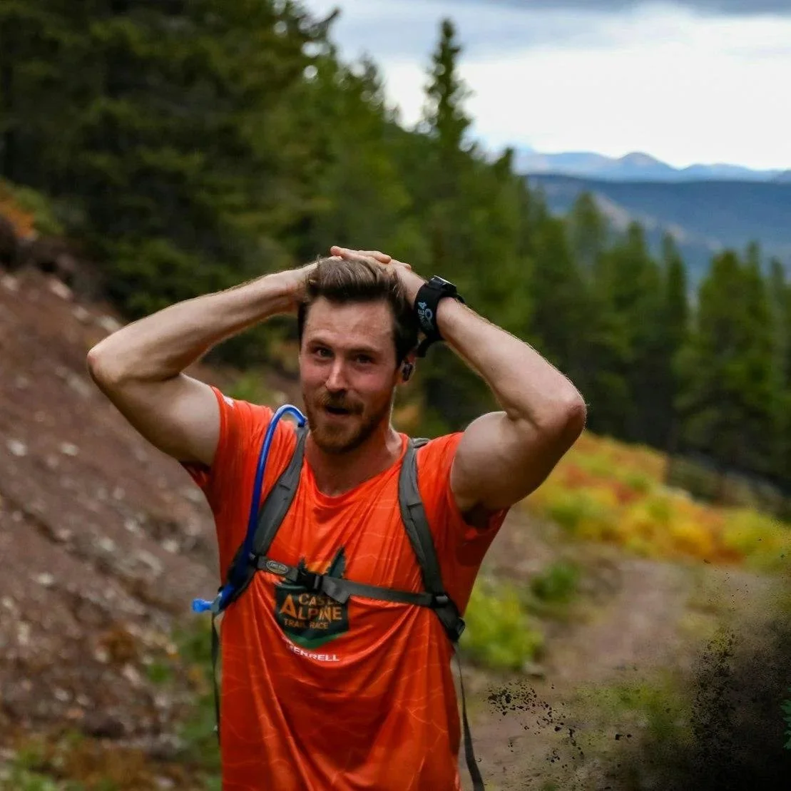 A man in an orange shirt with a backpack, standing outdoors on a trail, with mountains and trees in the background, appears to be after a run or hike.