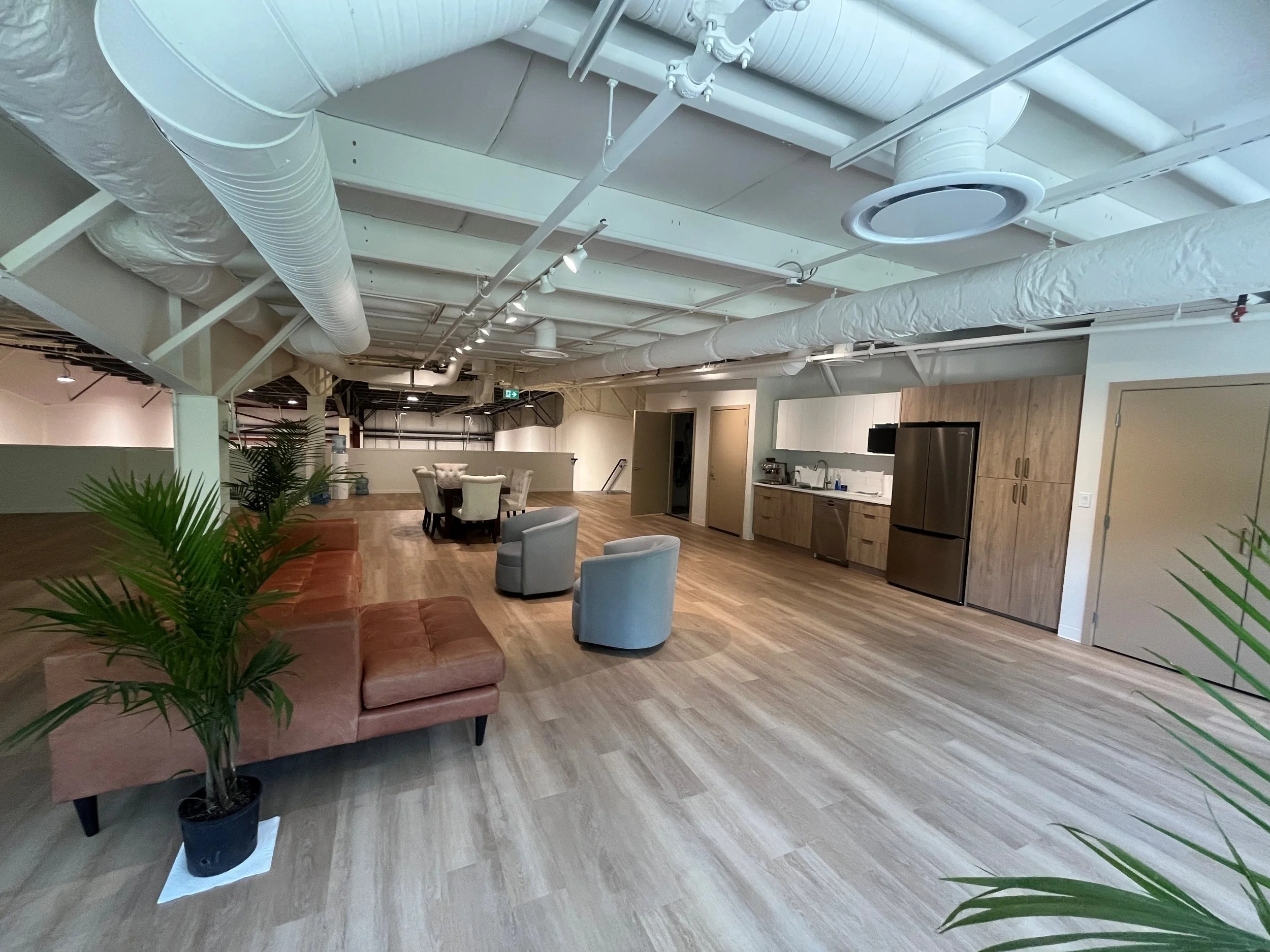 Indoor lounge area with sofas, chairs, and wooden cabinets, with white ductwork and pipes visible on the ceiling.