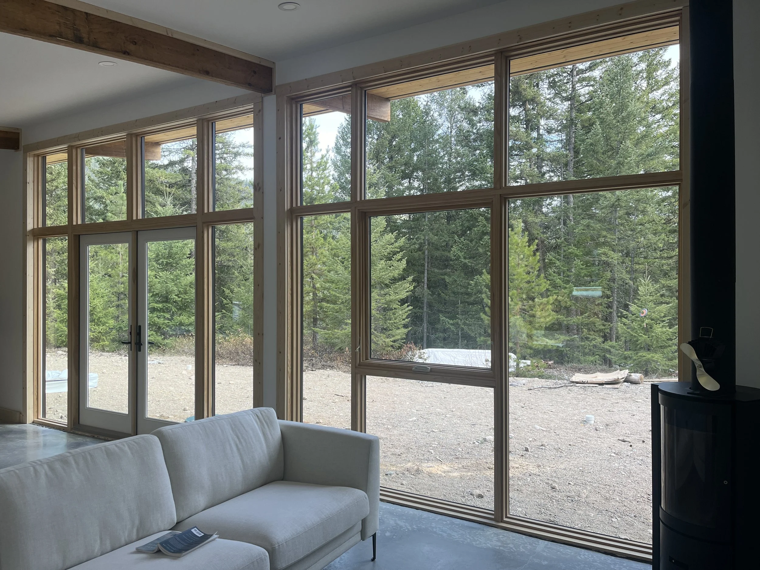 Interior view of a living room with large picture windows showing a forest outside, a white sofa, and a black stove in the corner.