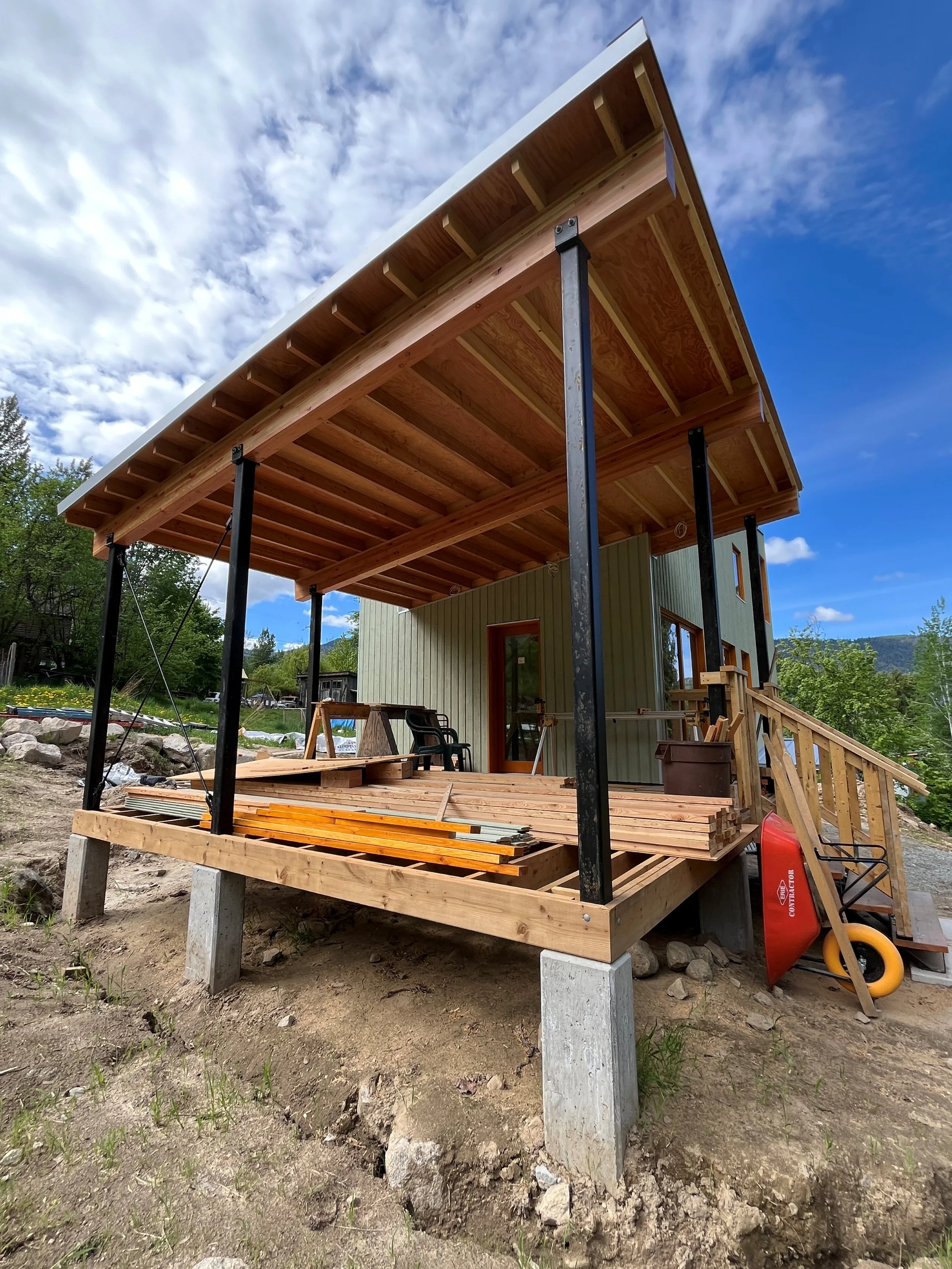 Under construction wooden deck with support posts and partially built roof on a new house, surrounded by construction tools and materials, with a cloudy sky and trees in the background.