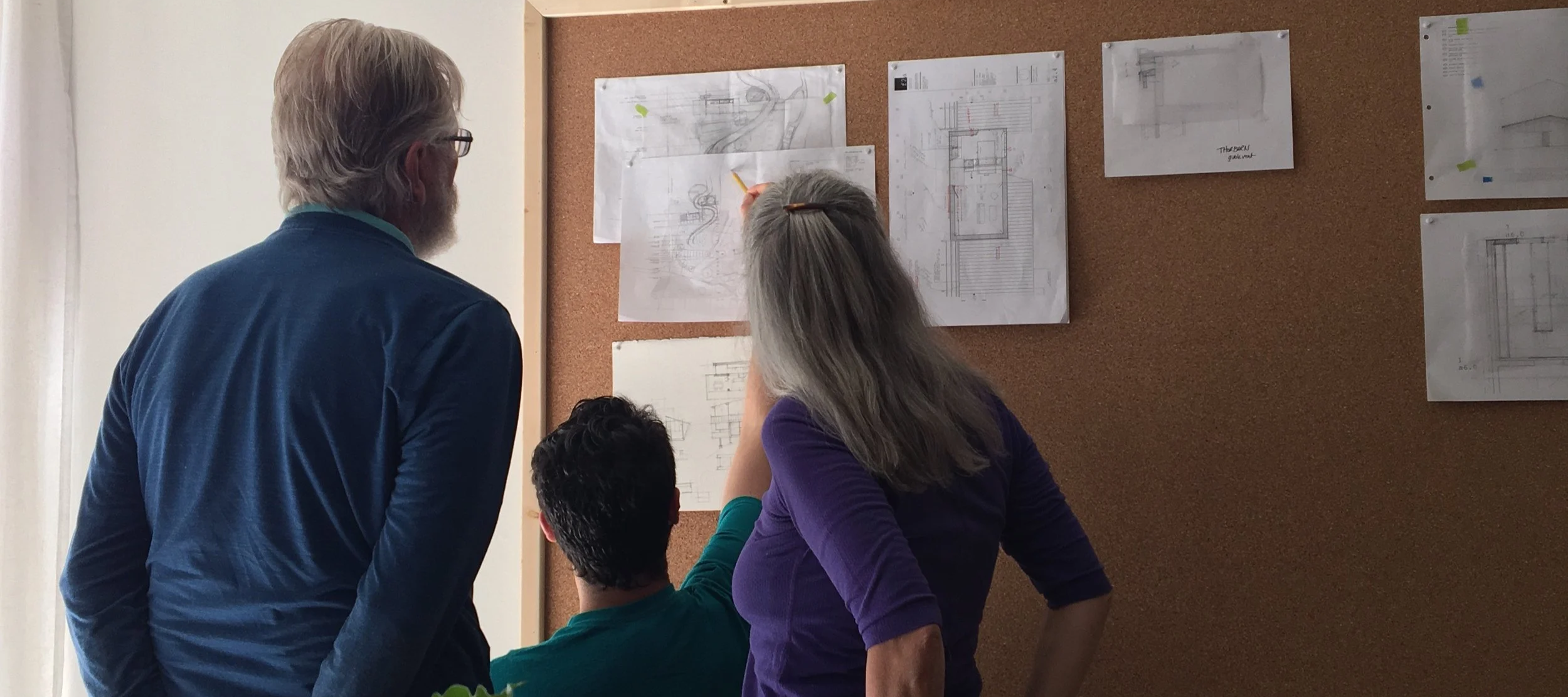 Three people discussing architectural plans pinned on a corkboard in an office or meeting room.
