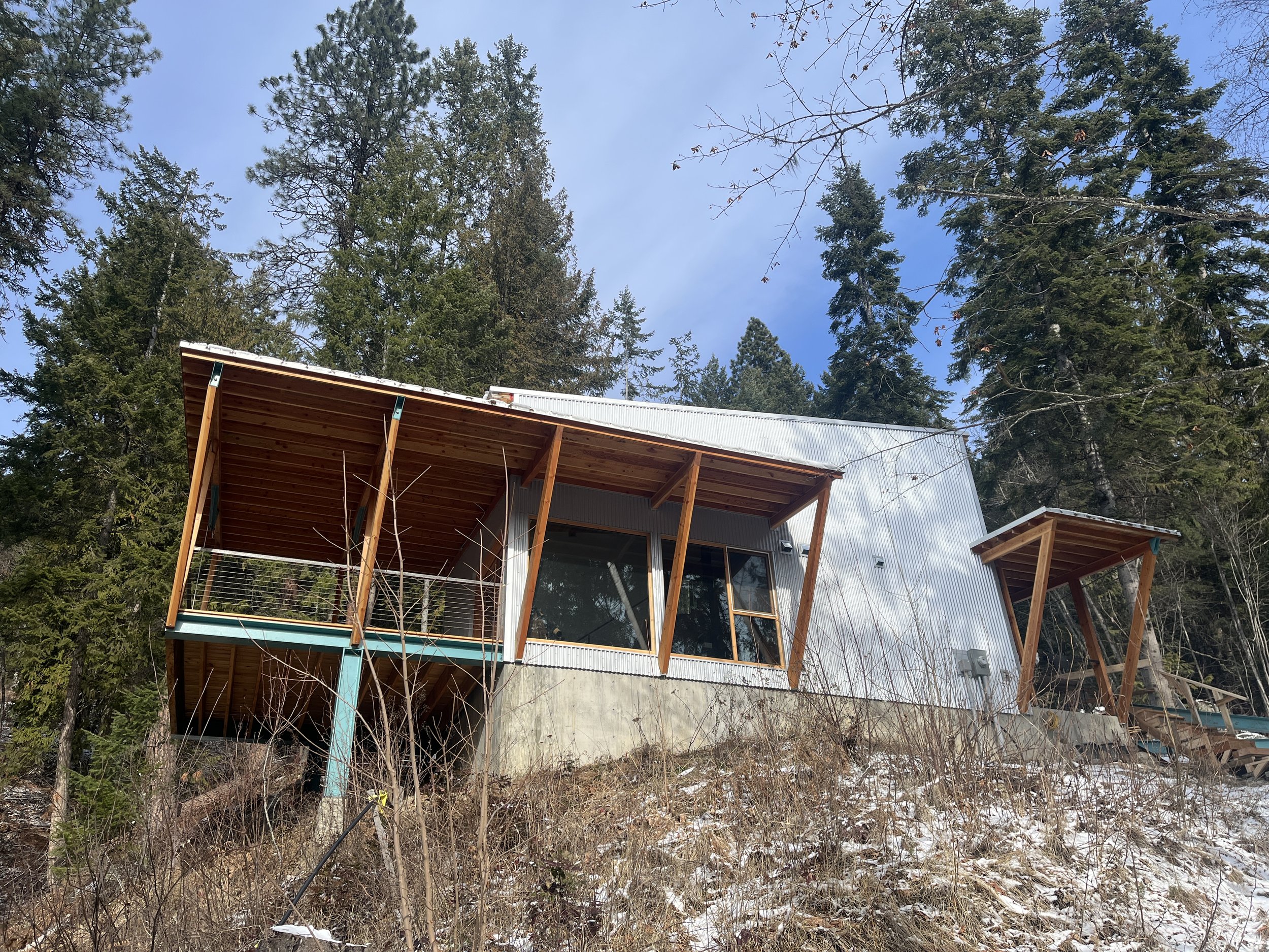Modern house with wooden balconies on a hillside surrounded by tall trees and dry grass with patches of snow, under a partly cloudy sky.