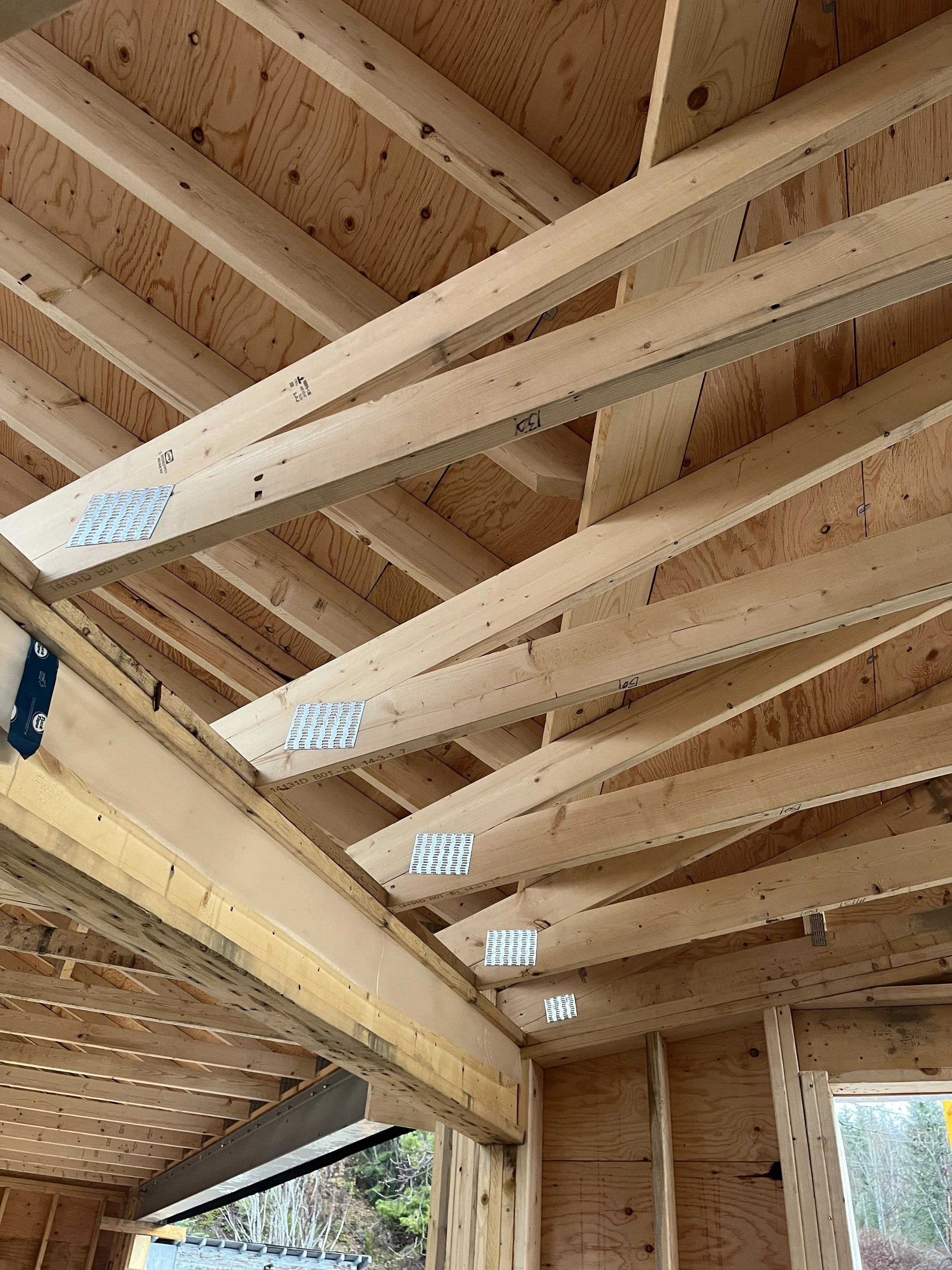 Interior view of a house under construction, showing exposed wooden framing, ceiling joists, and some metal brackets attaching the beams.