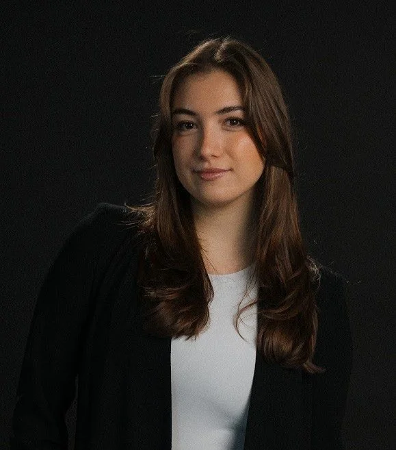 A young woman with long brown hair wearing a black blazer and white top, posing against a black background.