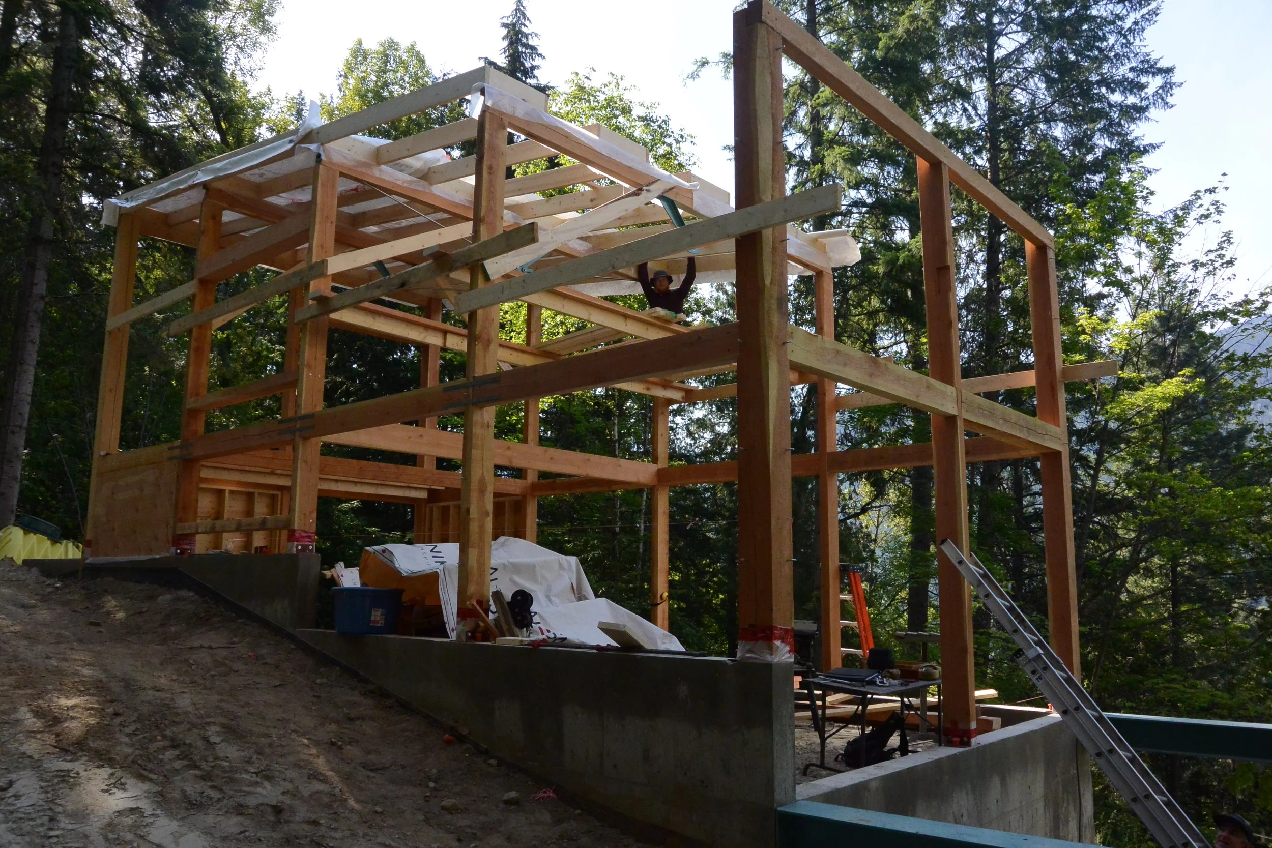 Construction site of a multi-story wooden house in a forested area with scaffolding, ladders, and building materials.