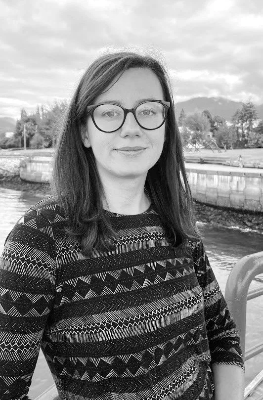 A woman with straight hair, glasses, and a patterned sweater standing outdoors near a river or canal, with trees, a stone wall, and mountains in the background.