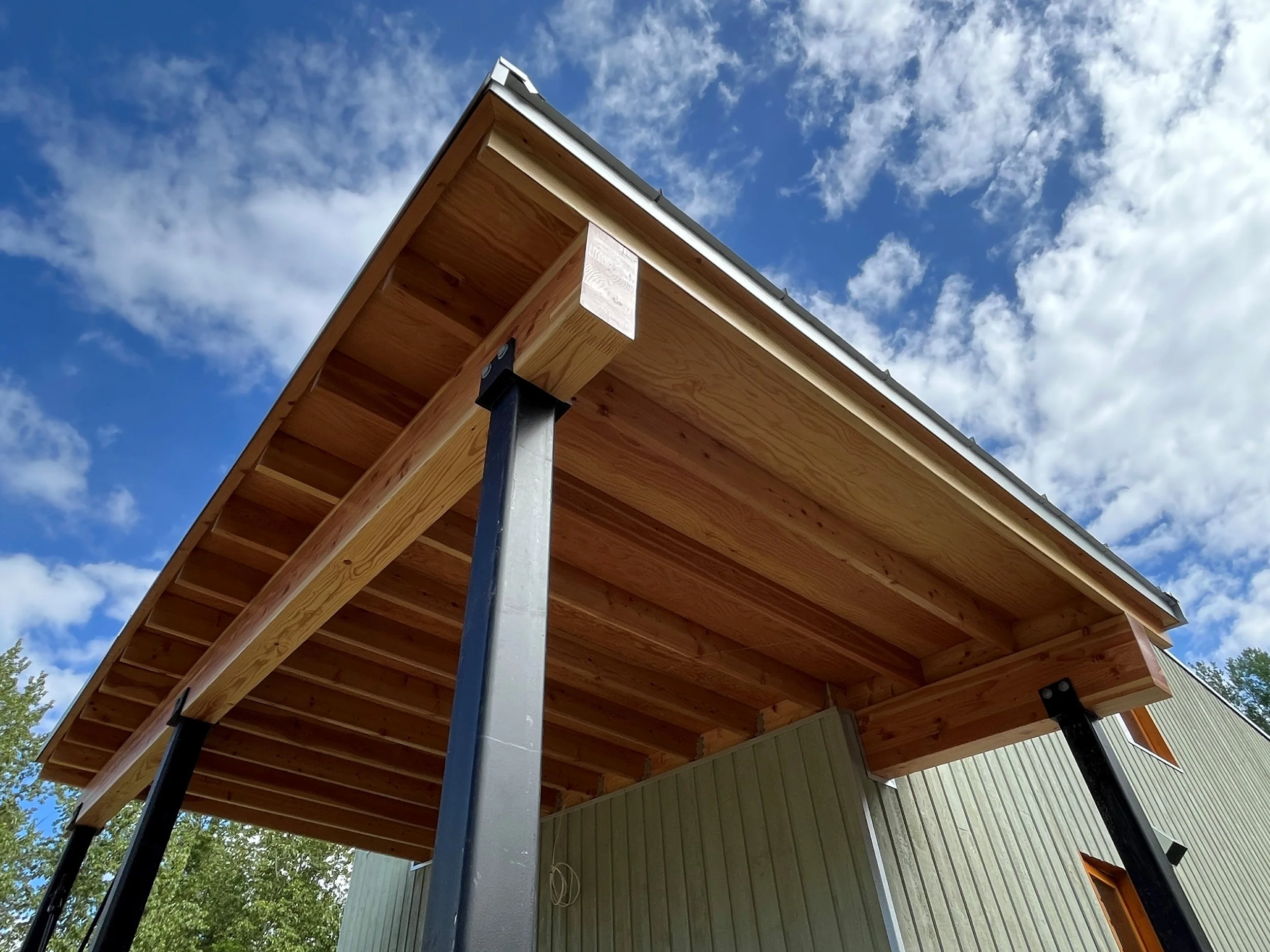 Underside of a modern house extension with a wooden roof, supported by black metal posts and tall support beams, against a blue sky with scattered clouds.