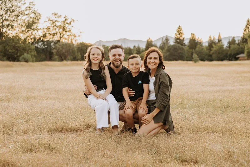 Family of four sitting on a grassy field at sunset, with mountains and trees in the background. The two children are sitting on the parents' laps, all smiling at the camera.