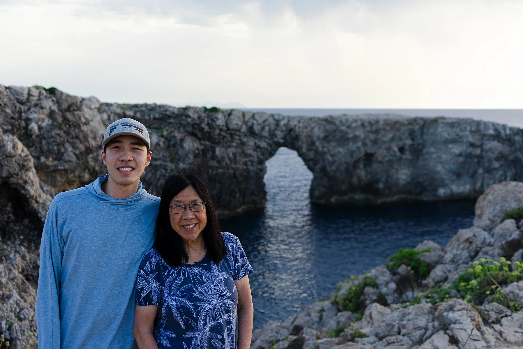 My mom and I (son) pose for a picture in front of an arch shaped rock on the coast of Menorca, Spain.