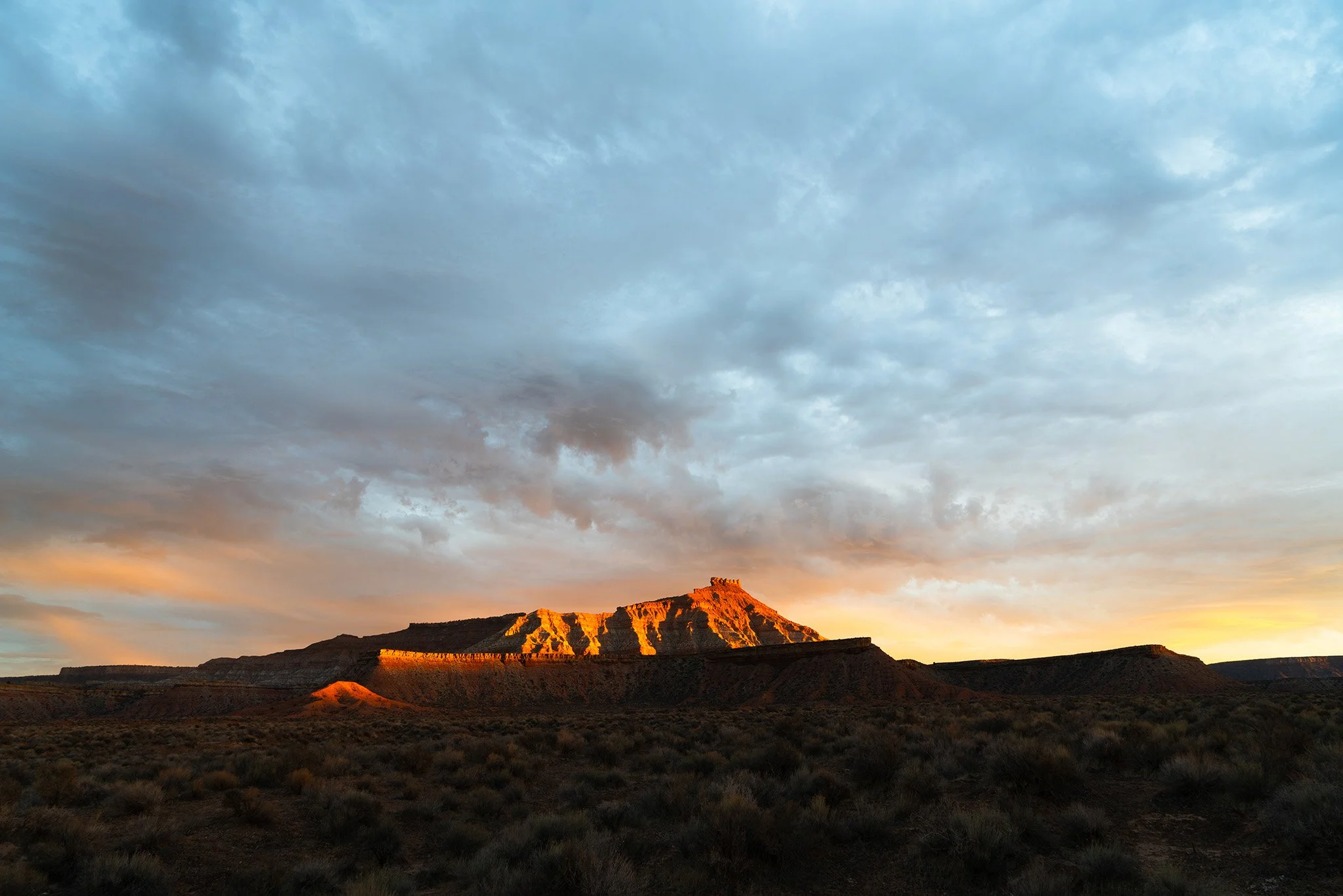 Yellow sunset colors reflect off of a butte in southern Utah with stormy clouds above and some shrubs in the foreground