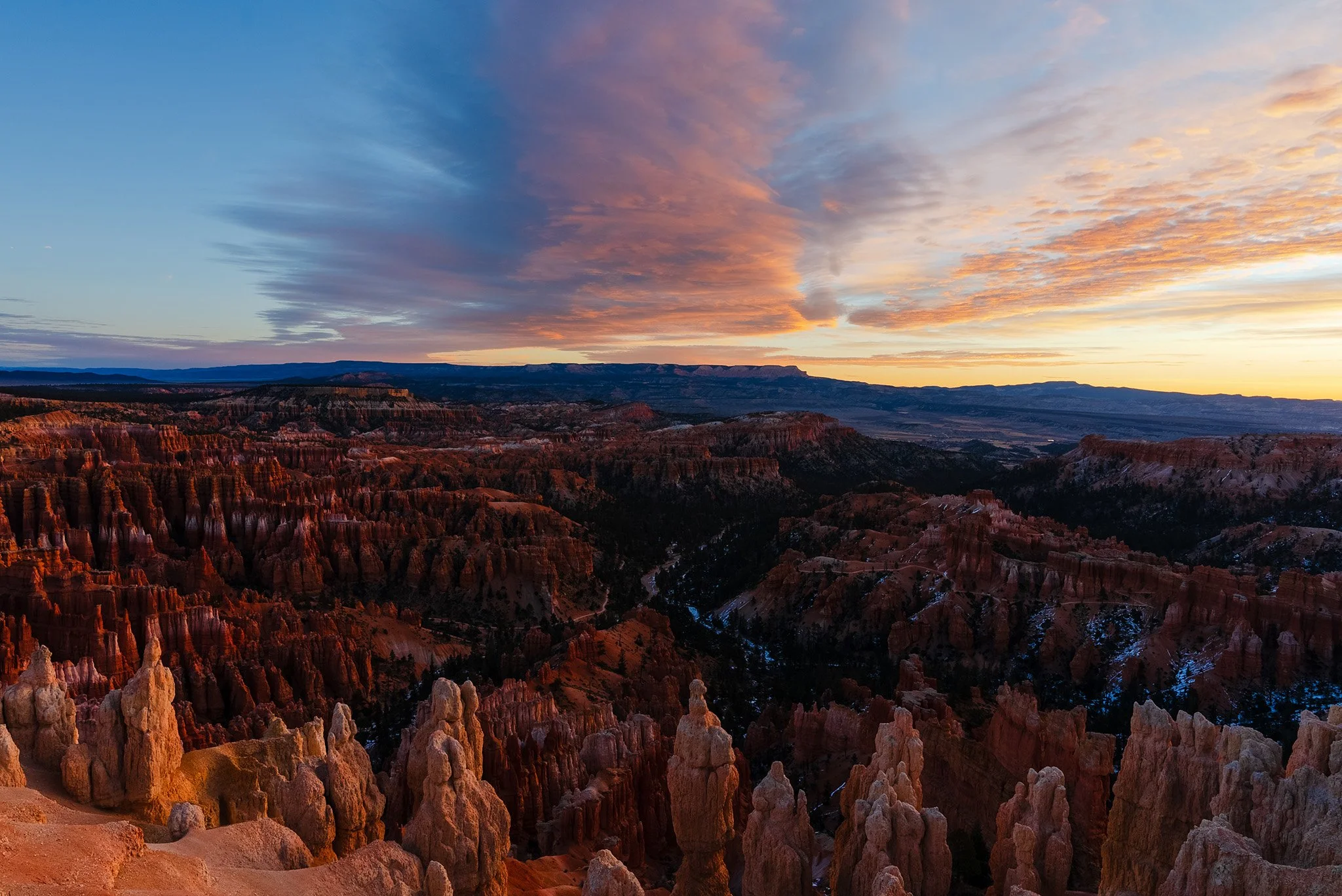 A wide-angle view of the Bryce Canyon National Park amphitheater during sunrise with some colorful clouds above and some hoodoos along the cliffs in the foreground.