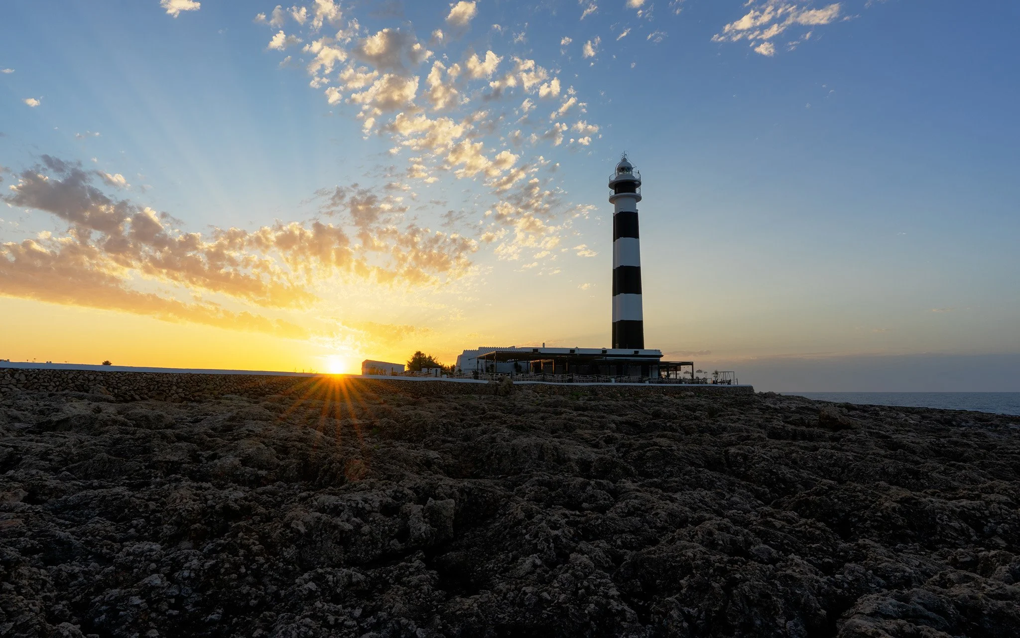 The Cap d'Artrutx Lighthouse at sunrise with a sunstar breaking the horizon and the light rays reflecting off the clouds above in Menorca, Spain.
