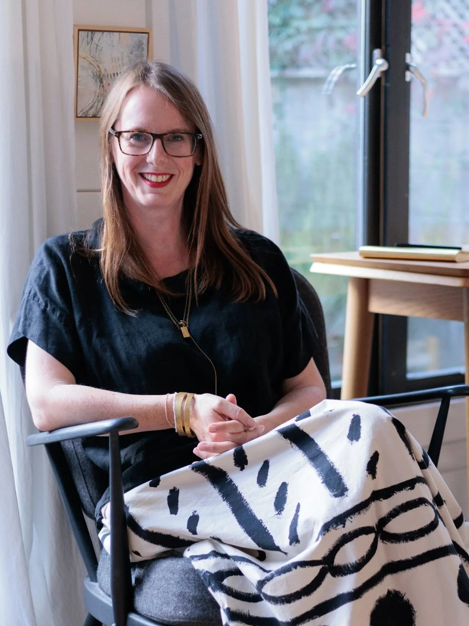 Michaela Murphy, therapist with glasses and long brown hair, wearing a black top, sitting in a chair, smiling. She is indoors in a therapy room near a window with a view of trees outside, and there are some books and a desk nearby.