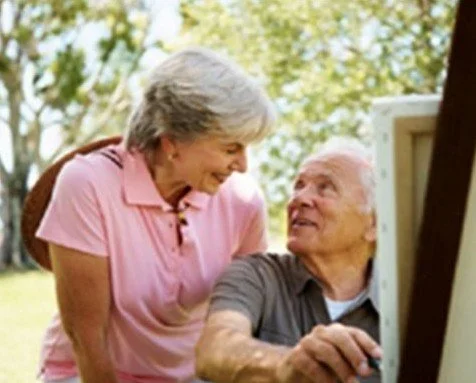 An elderly man and woman smiling and looking at each other outdoors near a computer or machine.