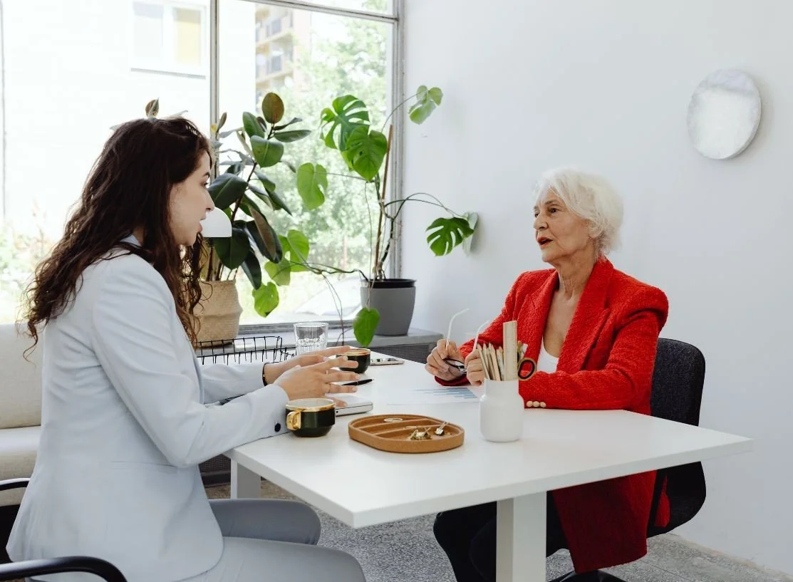 A young female doctor and an elderly woman are sitting at a white table in a bright room with large windows and green plants. The doctor is holding a black mug, and the elderly woman is holding a pair of glasses. There are various items on the table, including a white vase with books or magazines, a wooden tray with jewelry, and a glass of water. The elderly woman is wearing a red blazer, and the doctor is dressed in a white coat.