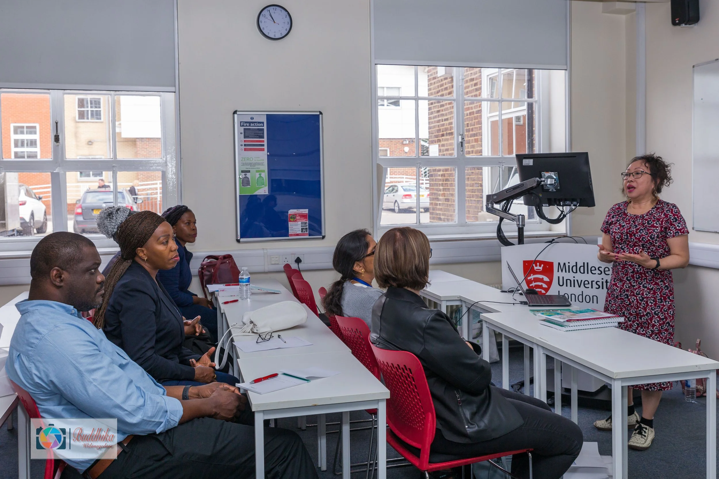 A classroom setting at Middlesex University London with a group of diverse students attending a lecture. A woman lecturer is standing near a desk with the university's logo, speaking to the students, who are seated at tables with notebooks and water bottles. Large windows and a wall clock are visible in the background.