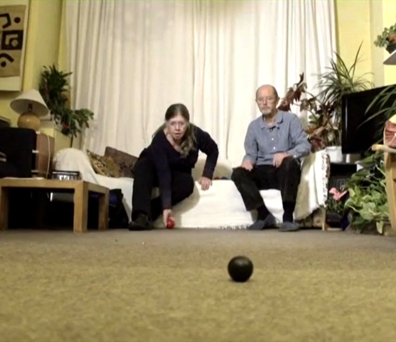 A living room with two people, a woman and a man, playing bocce ball on a carpeted floor. The woman is crouching down, holding a bocce ball, while the man sits on a sofa behind her. The room has a yellow wall, a window with curtains, houseplants, and furniture including a table with a lamp and a TV.