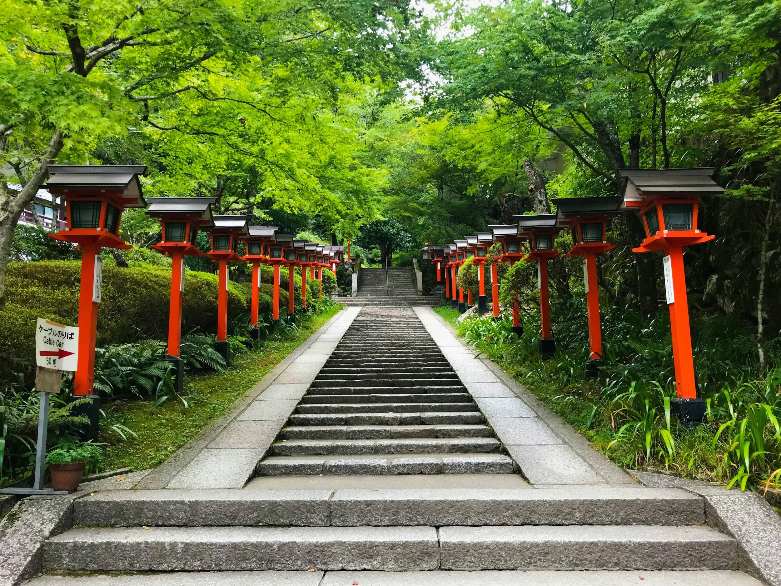 Stone stairway leading up through a path lined with traditional red Japanese lanterns on each side, surrounded by lush green trees and foliage.