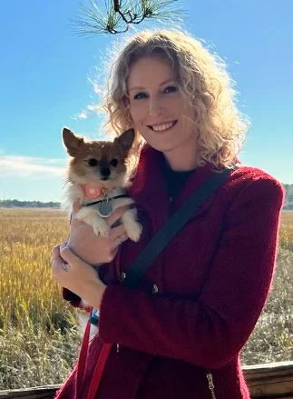A woman with curly blonde hair smiling while holding a small dog, with a field and blue sky in the background.