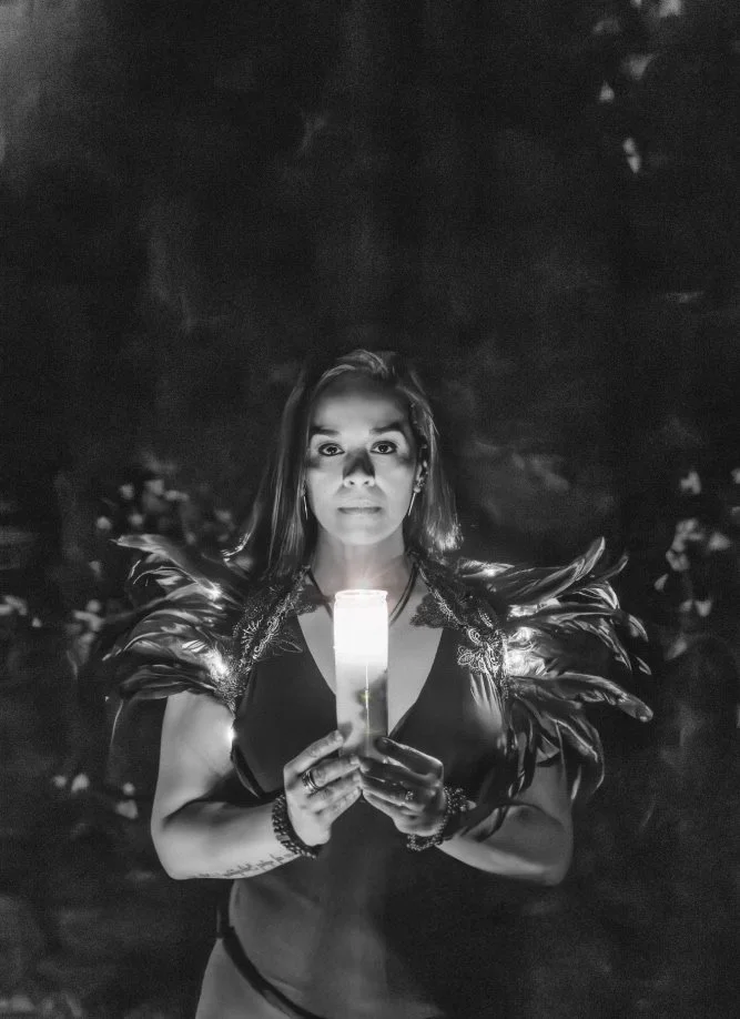 A woman holding a lit candle outdoors at night, dressed in a costume with feathered shoulder accessories.
