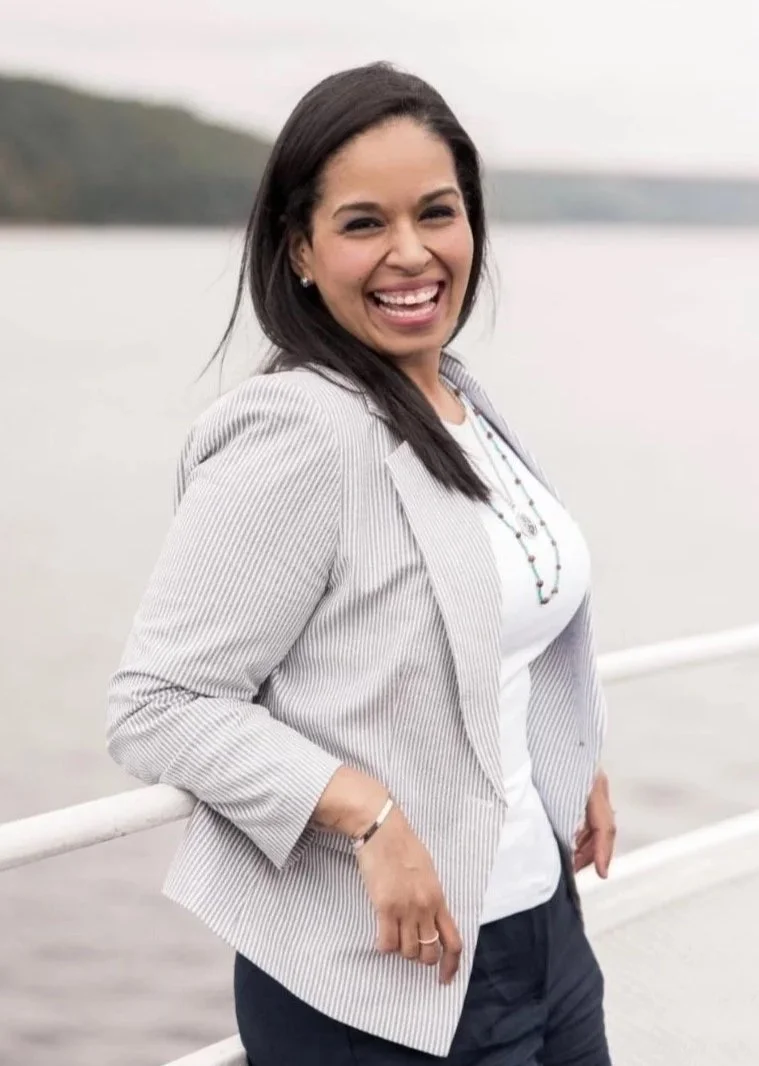 Woman smiling and posing outdoors near a body of water, wearing a light gray blazer and a white shirt.