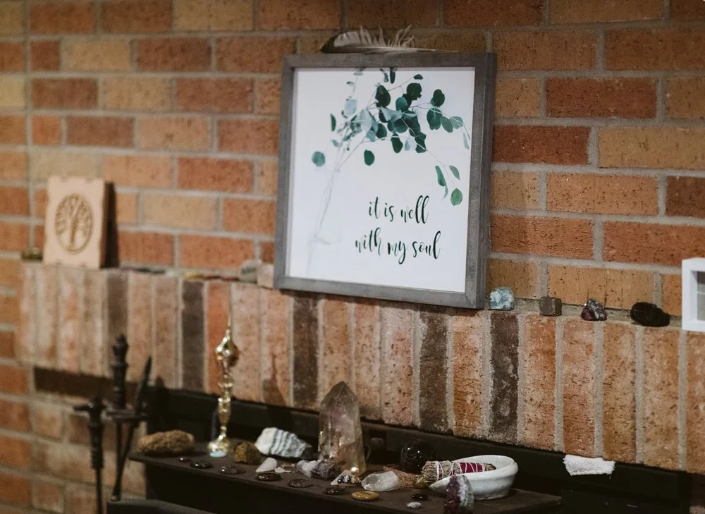A decorative display on a black shelf in front of a brick wall features crystals, stones, and a small white bowl filled with rocks. Above, a framed sign with a gray border displays green leaves and the quote, 'it is well with my soul'.