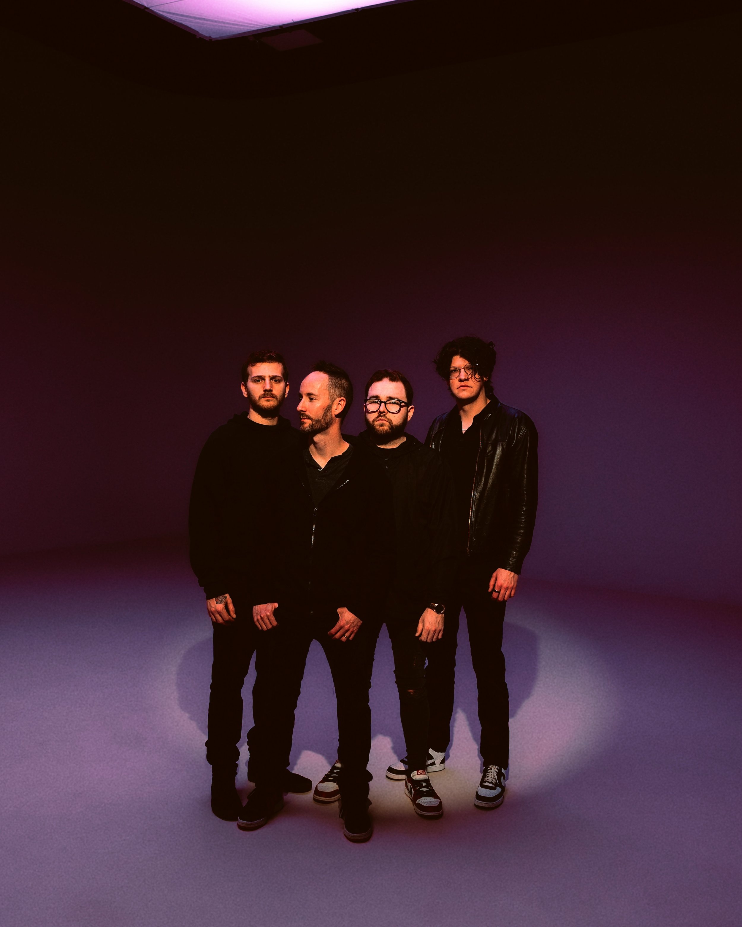 Four young men standing in front of a purple-lit wall. They have serious expressions, with one looking to the side, one with sunglasses, and two looking directly at the camera. All are dressed in dark casual clothing.