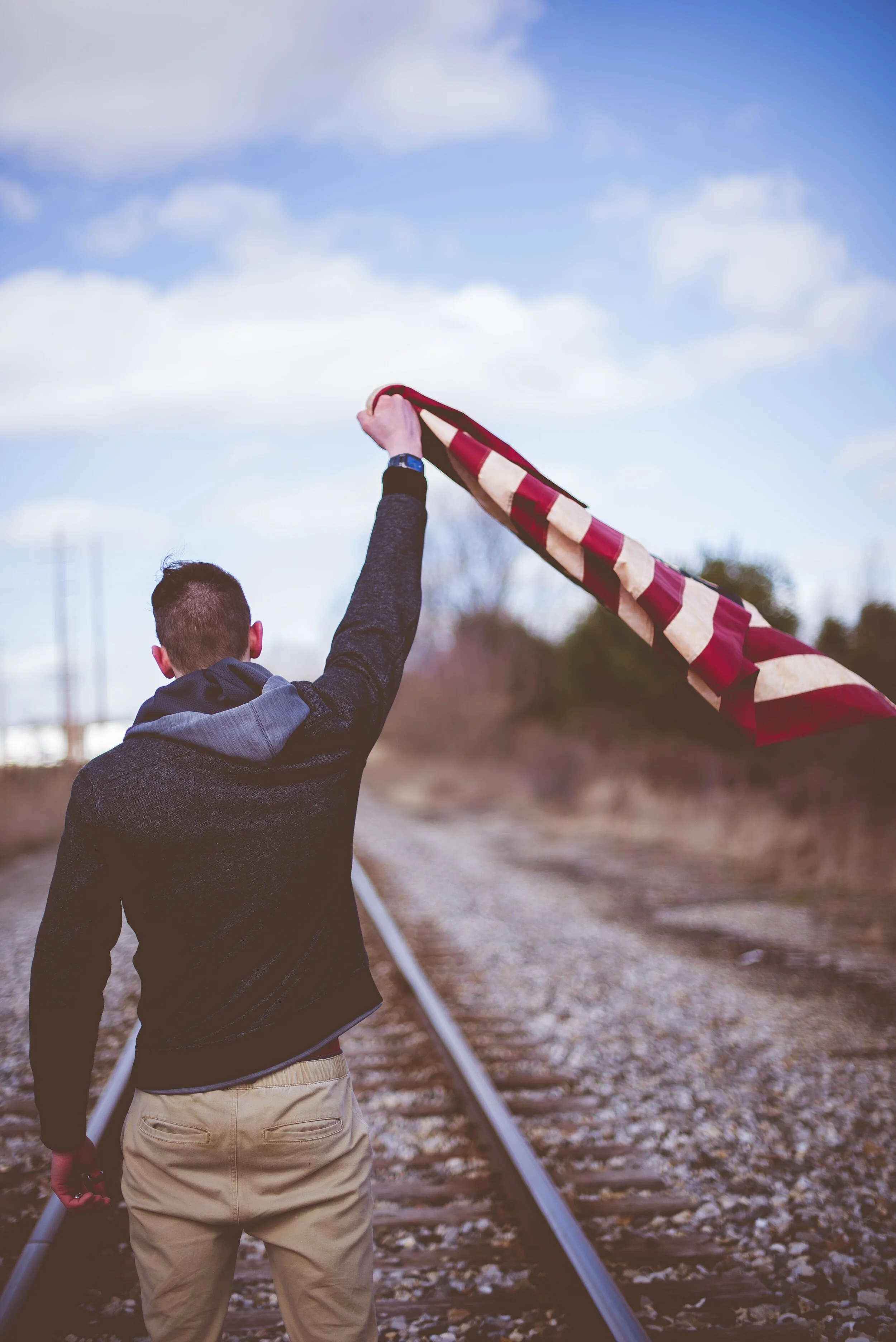 vertical-shot-male-standing-train-tracks-while-holding-up-united-states-flag.jpg