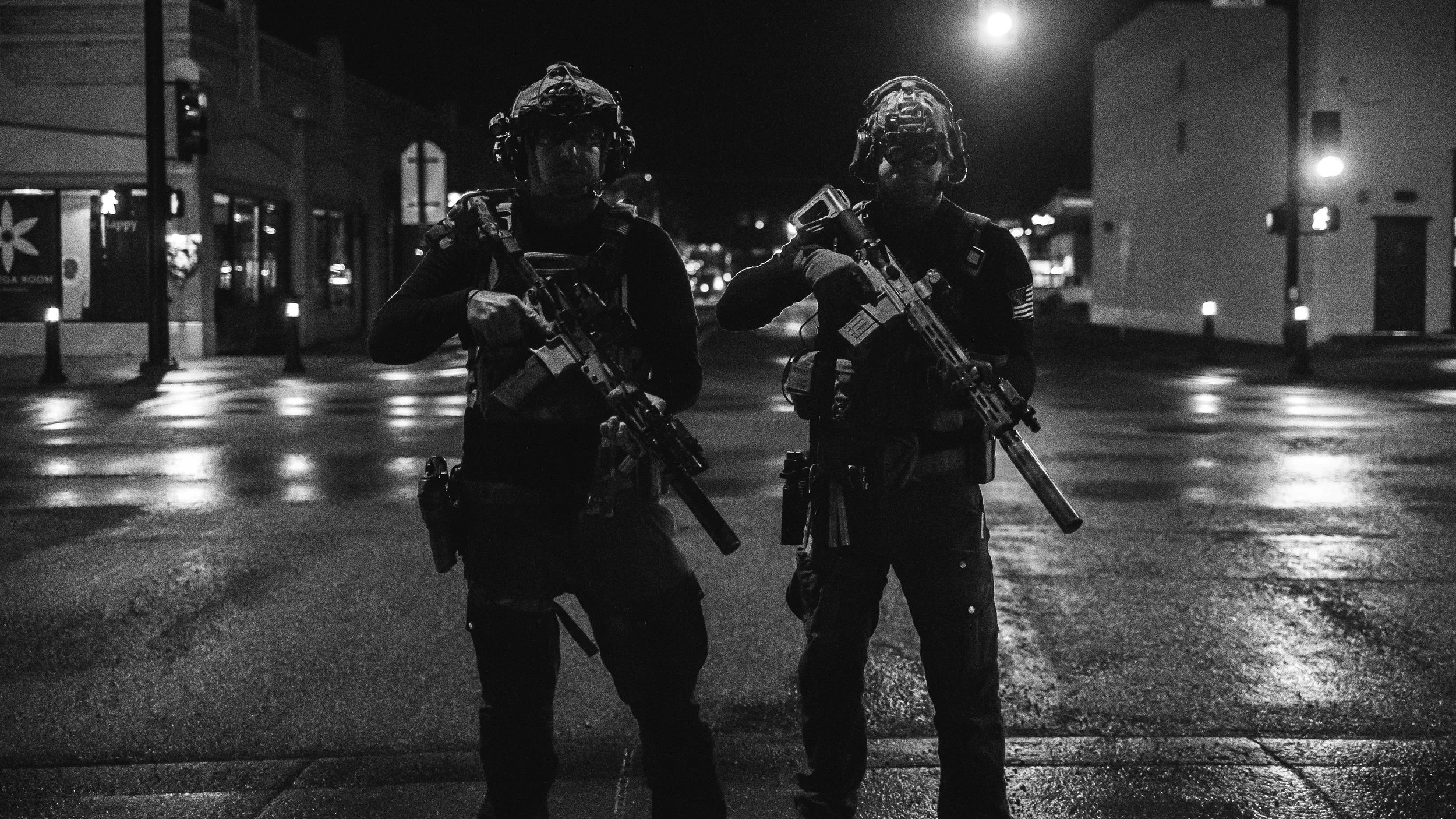 Two armed police officers wearing helmets and tactical gear standing on a wet city street at night.