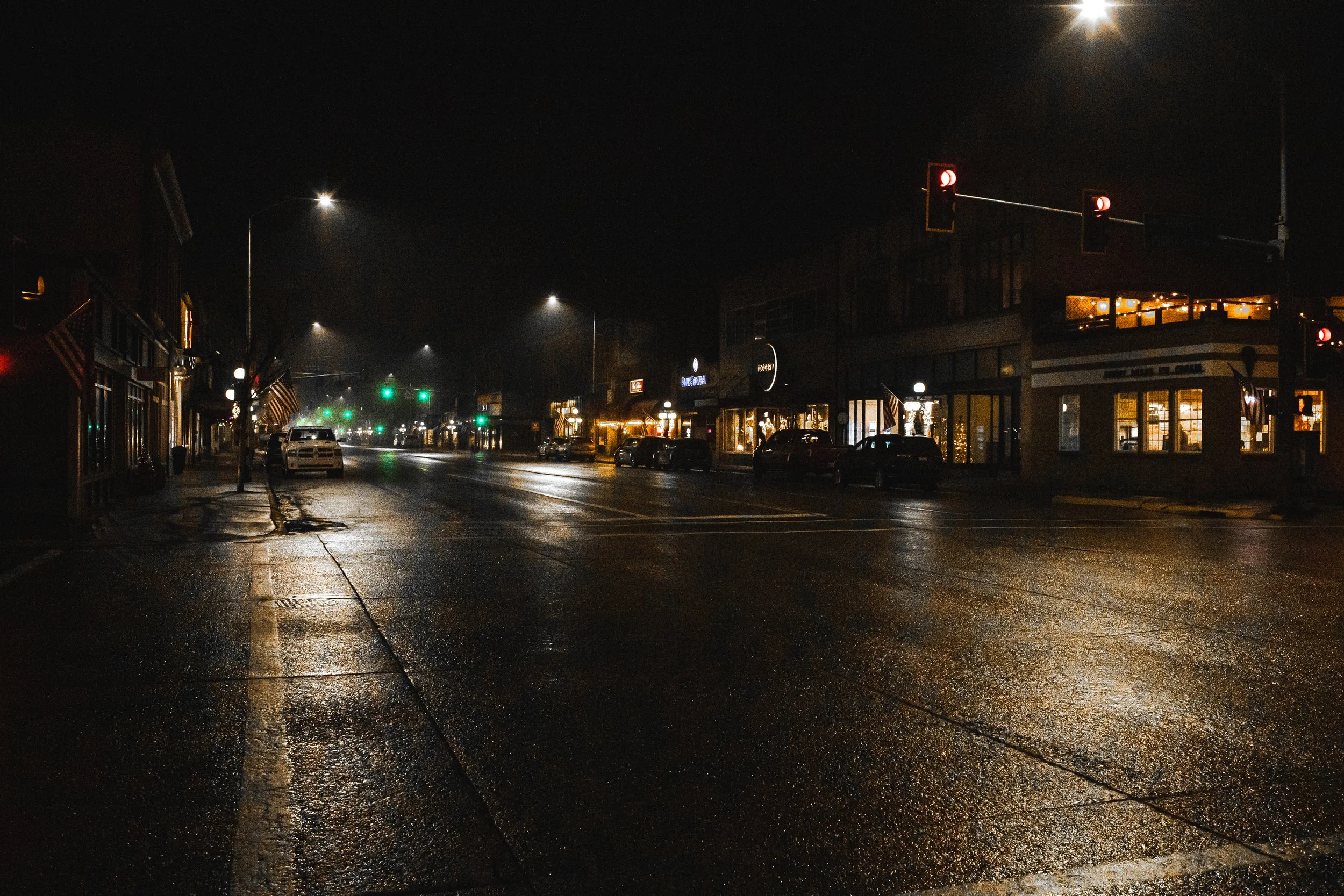 A quiet, wet city street at night with illuminated storefronts, parked cars, and traffic lights, some showing red. The street appears to be freshly rain-soaked, reflecting the streetlights and shop windows.