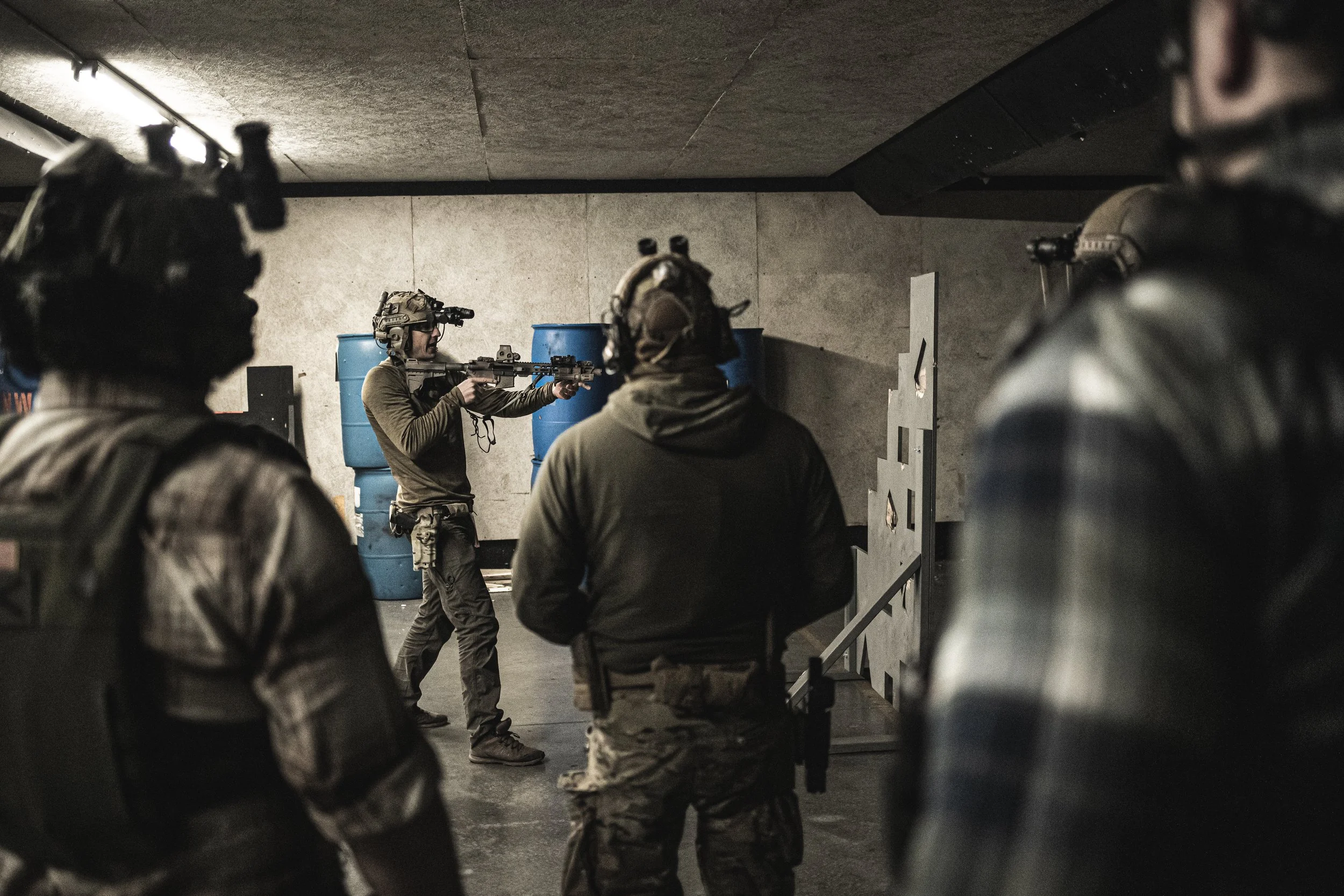Soldiers in military gear participating in a firearms training session indoors, with one soldier aiming and shooting a rifle while others observe.