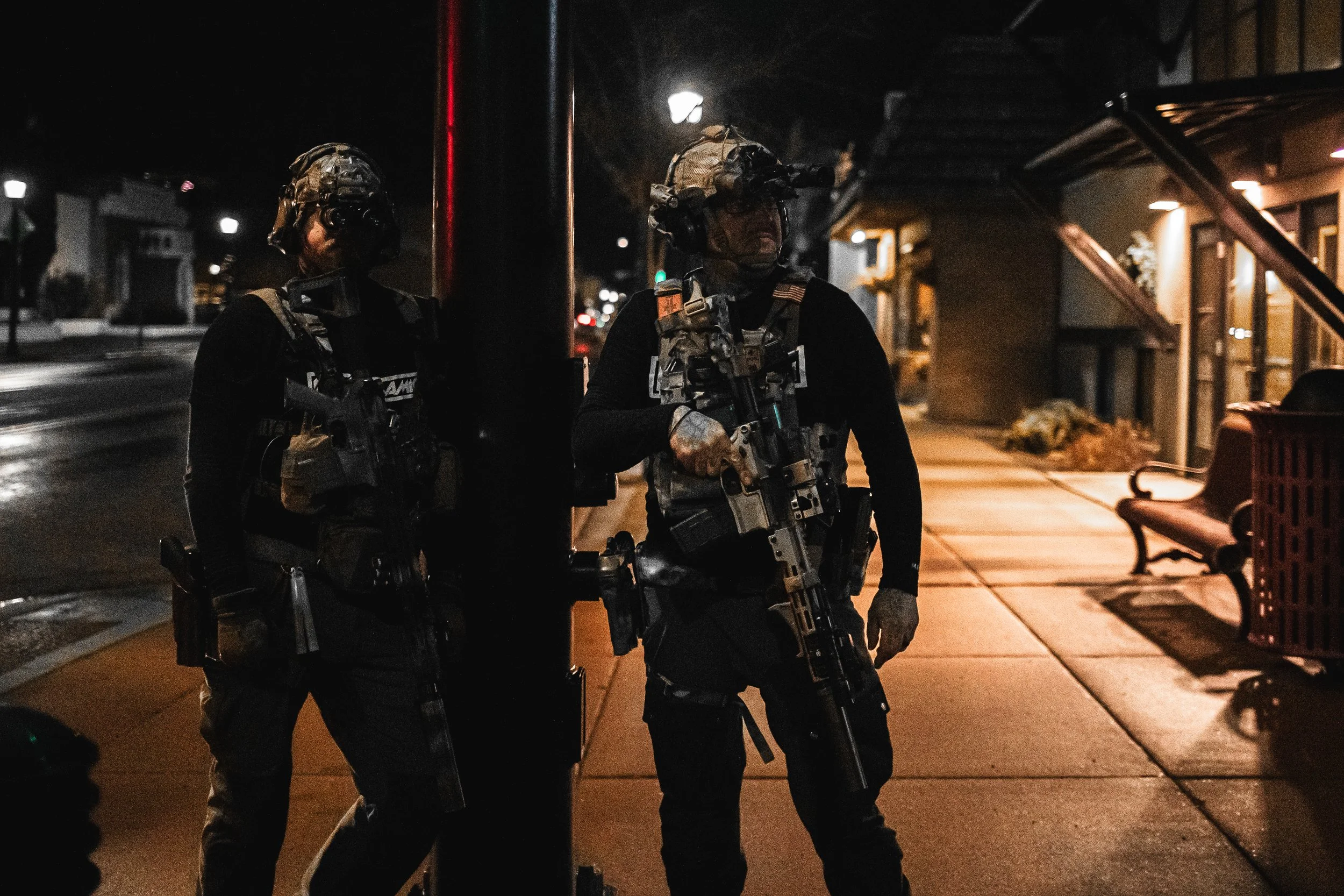 Two armed police officers in tactical gear standing guard on a city sidewalk at night, with nearby benches and buildings.