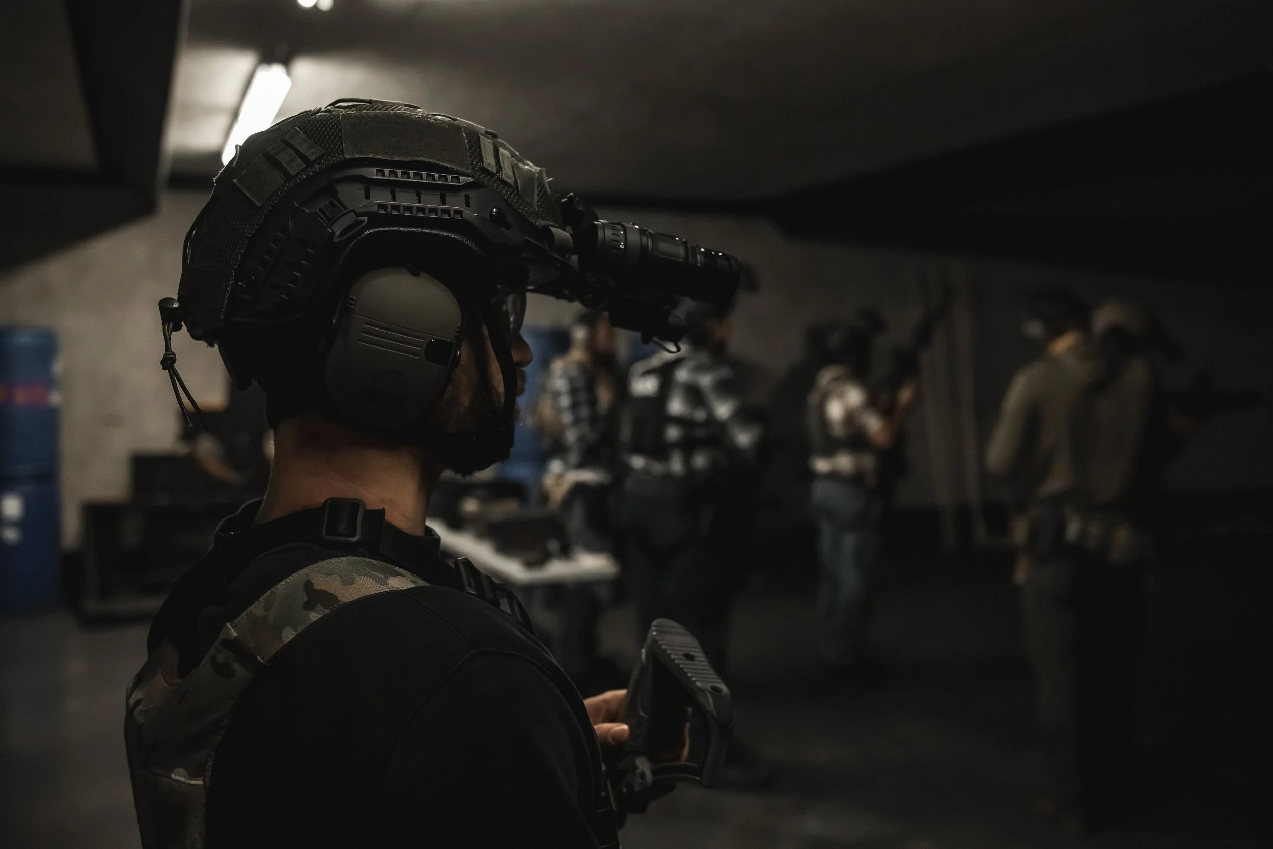 A man in military gear with a helmet and headset holding a rifle, standing in a dark room with other people in the background.