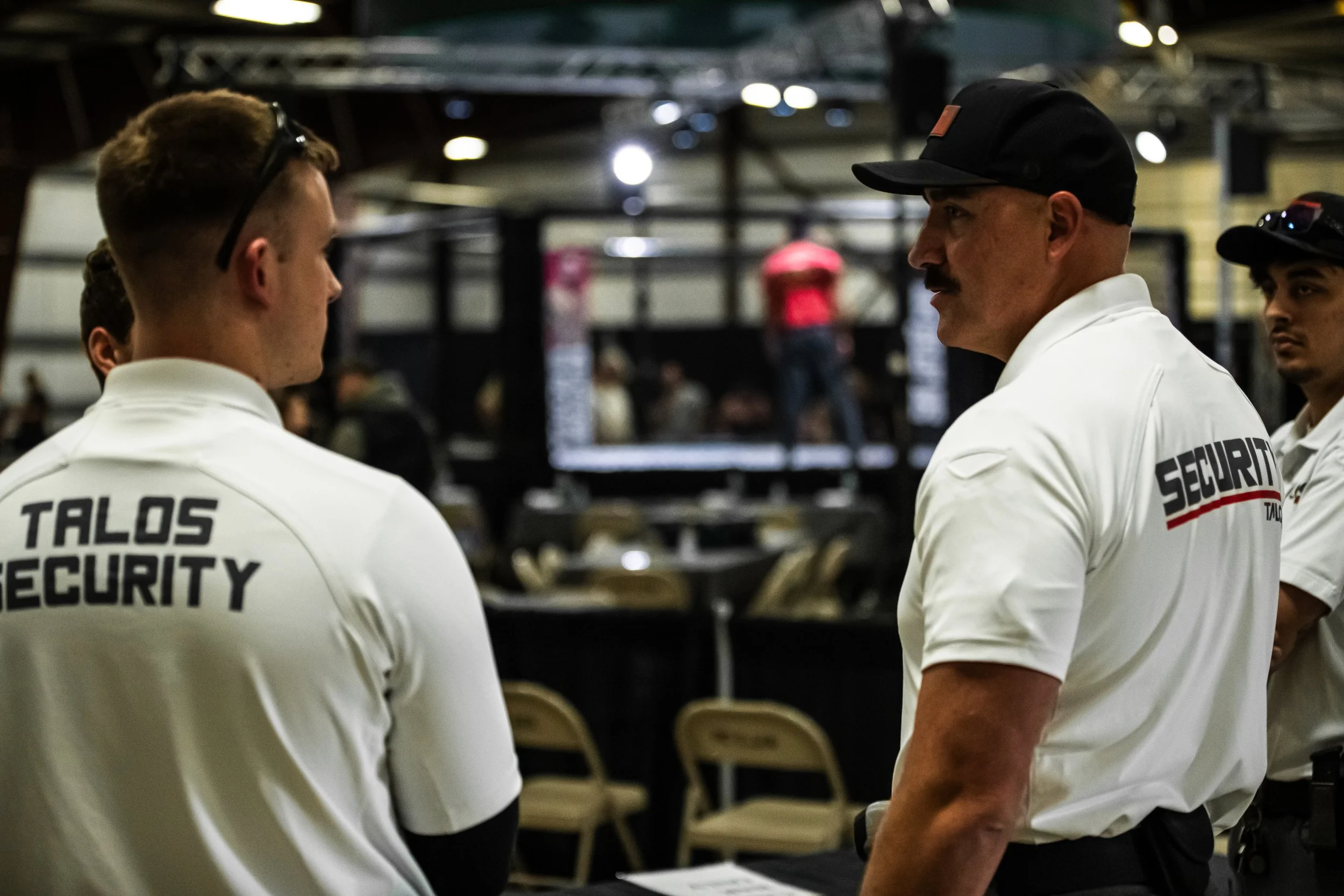 Security personnel wearing white shirts with 'TALOS SECURITY' and 'SECURITY' printed on the back, standing and talking in an indoor event space with a dark background and chairs.