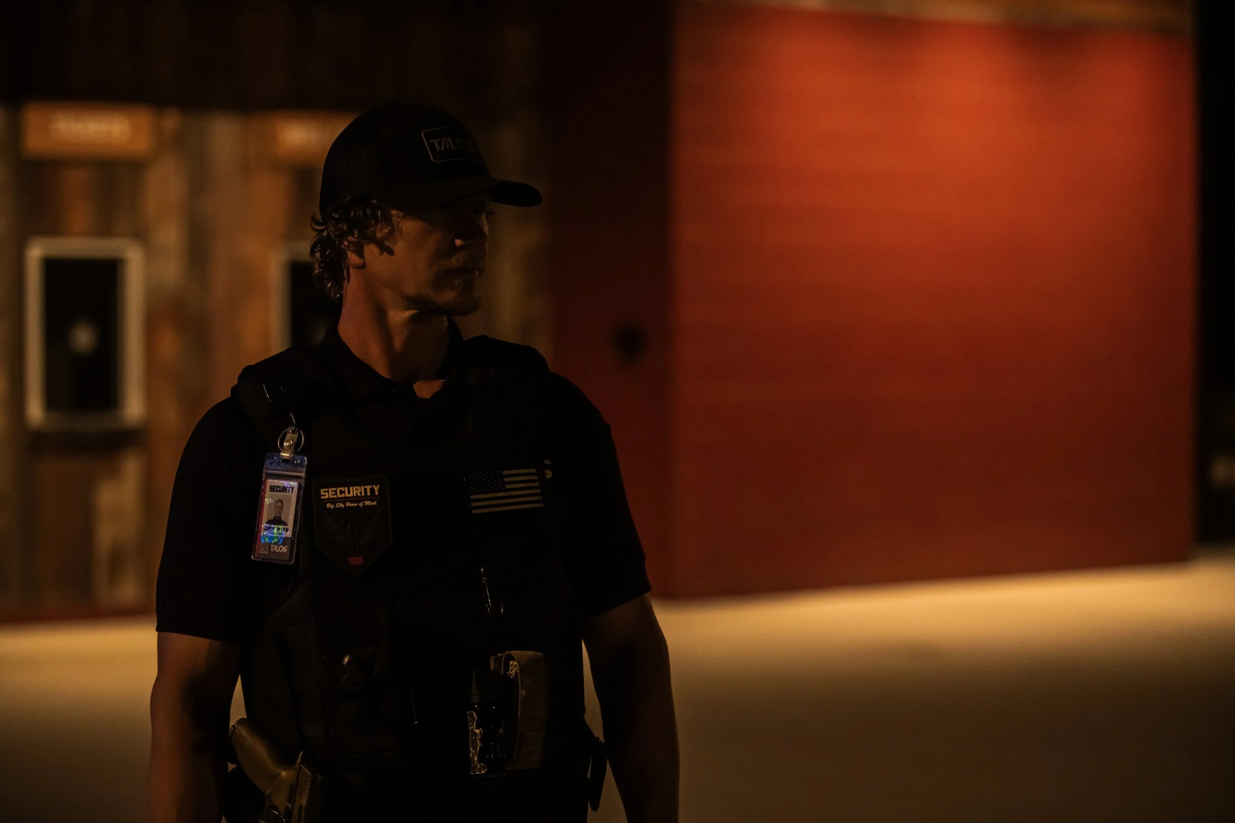A security guard standing outdoors at night, wearing a black uniform and cap, with an ID badge and patches on his shirt, near a building with a wooden wall and a red rectangular wall in the background.