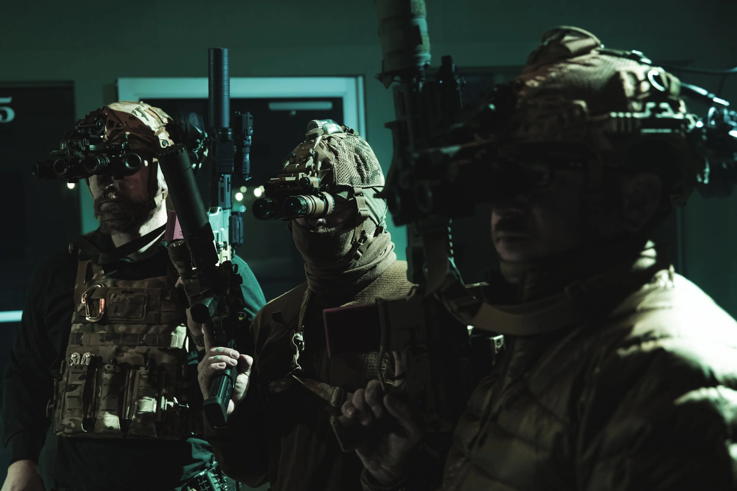 Three soldiers in tactical gear with night vision goggles in a dark, indoor setting.