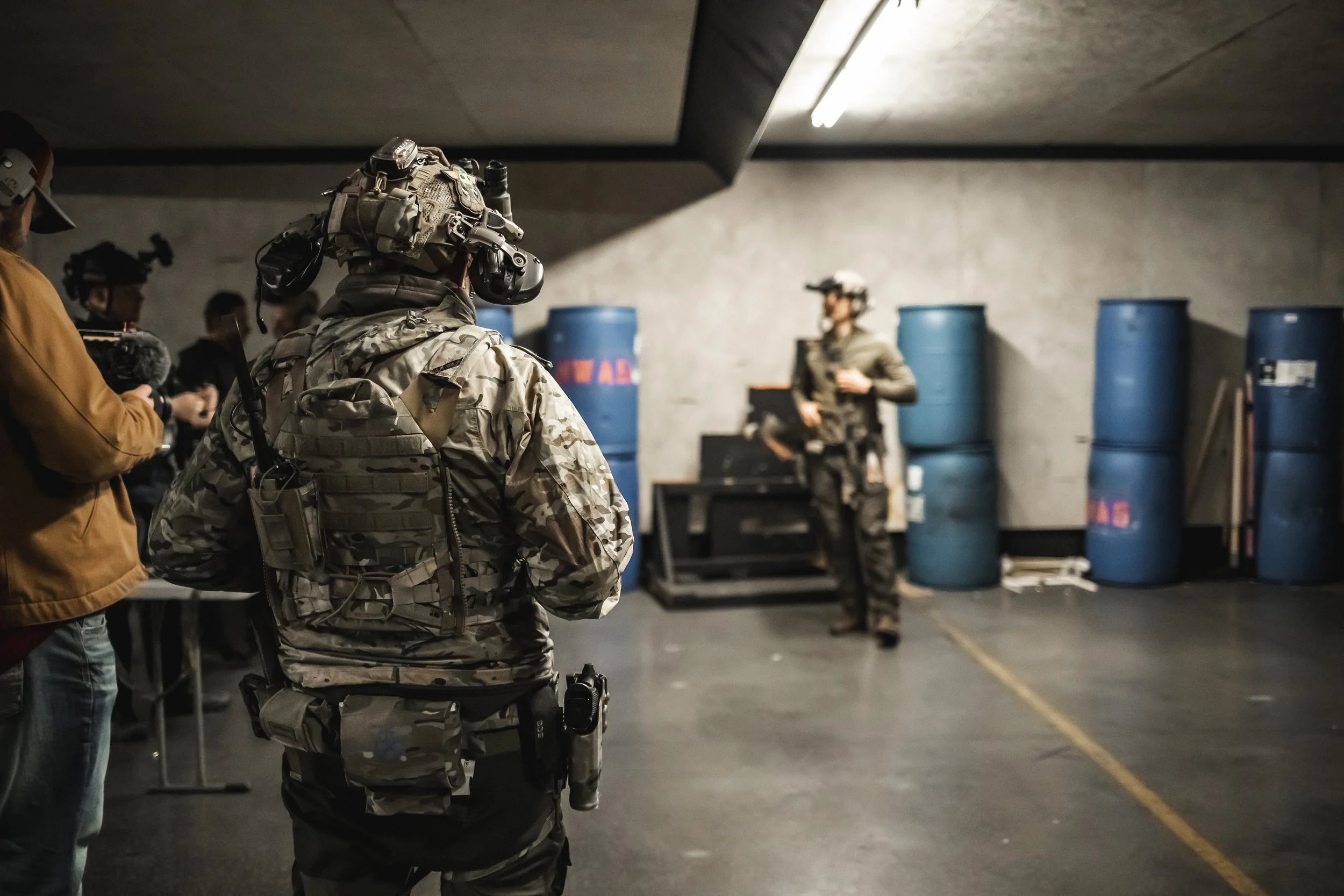 Military personnel in camouflage uniforms and tactical gear standing in a training or briefing room with blue barrels and equipment in the background.