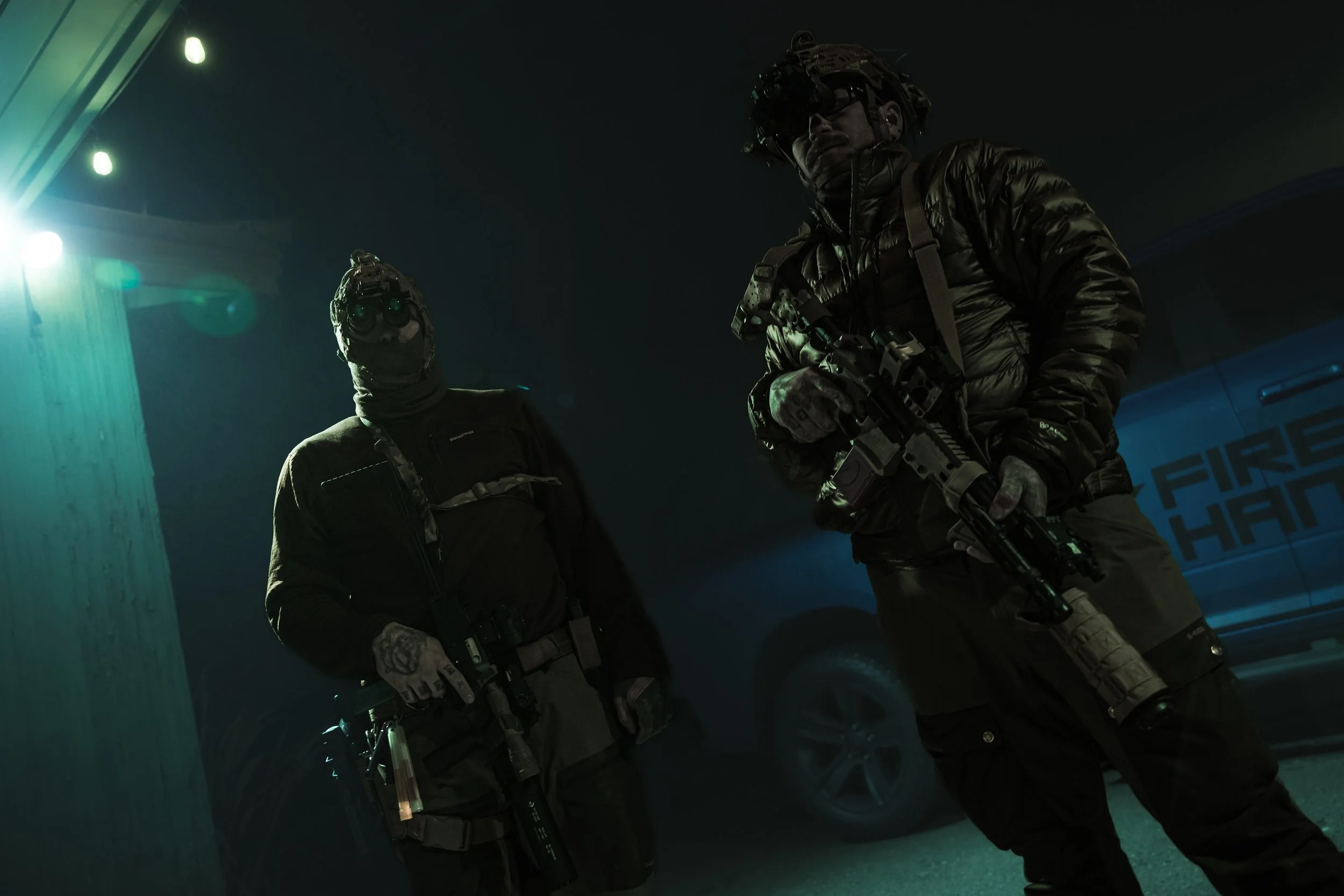 Two armed tactical officers in dark clothing and gear stand in low light near a police vehicle at night.