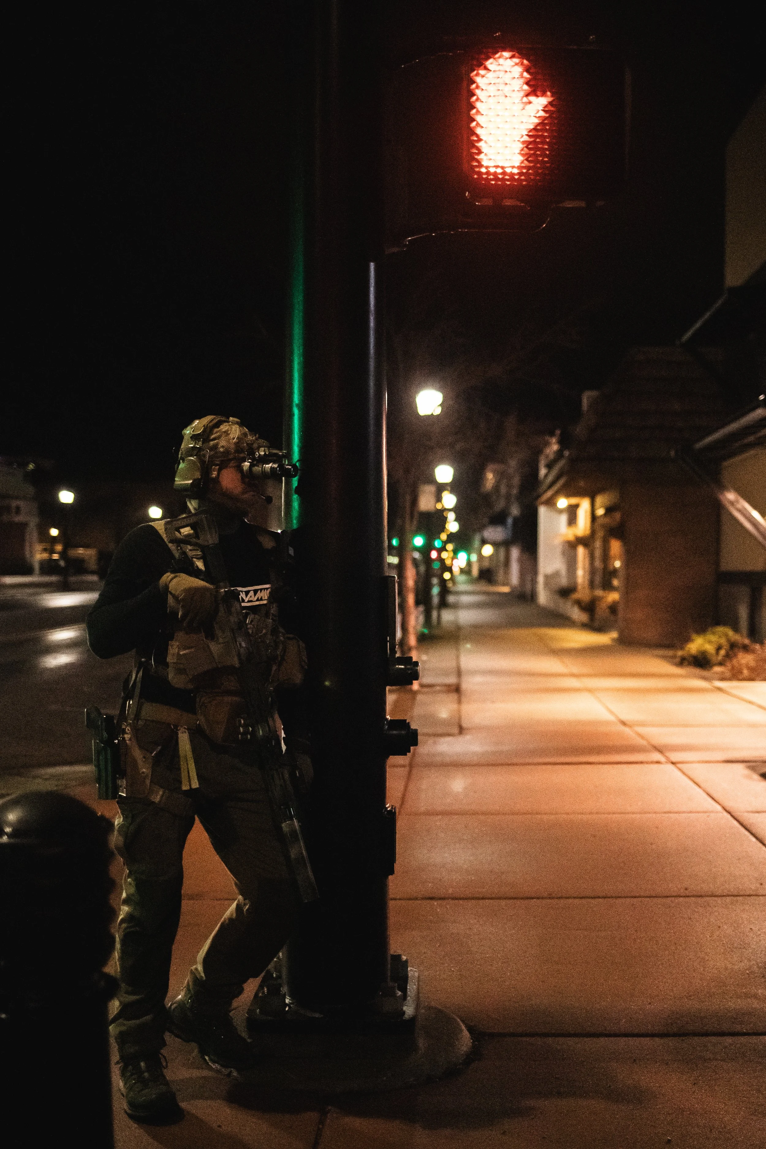 A armed soldier standing on a sidewalk at night near a red pedestrian signal.